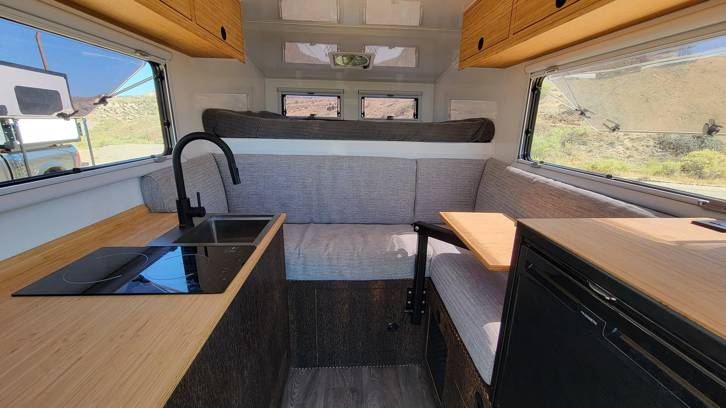 Interior of a compact camper van with a small kitchen area, including a black induction cooktop, a sink with a black faucet, and wooden countertops. There is a gray cushioned seating area with a table, and a sleeping area with a mattress at the back.