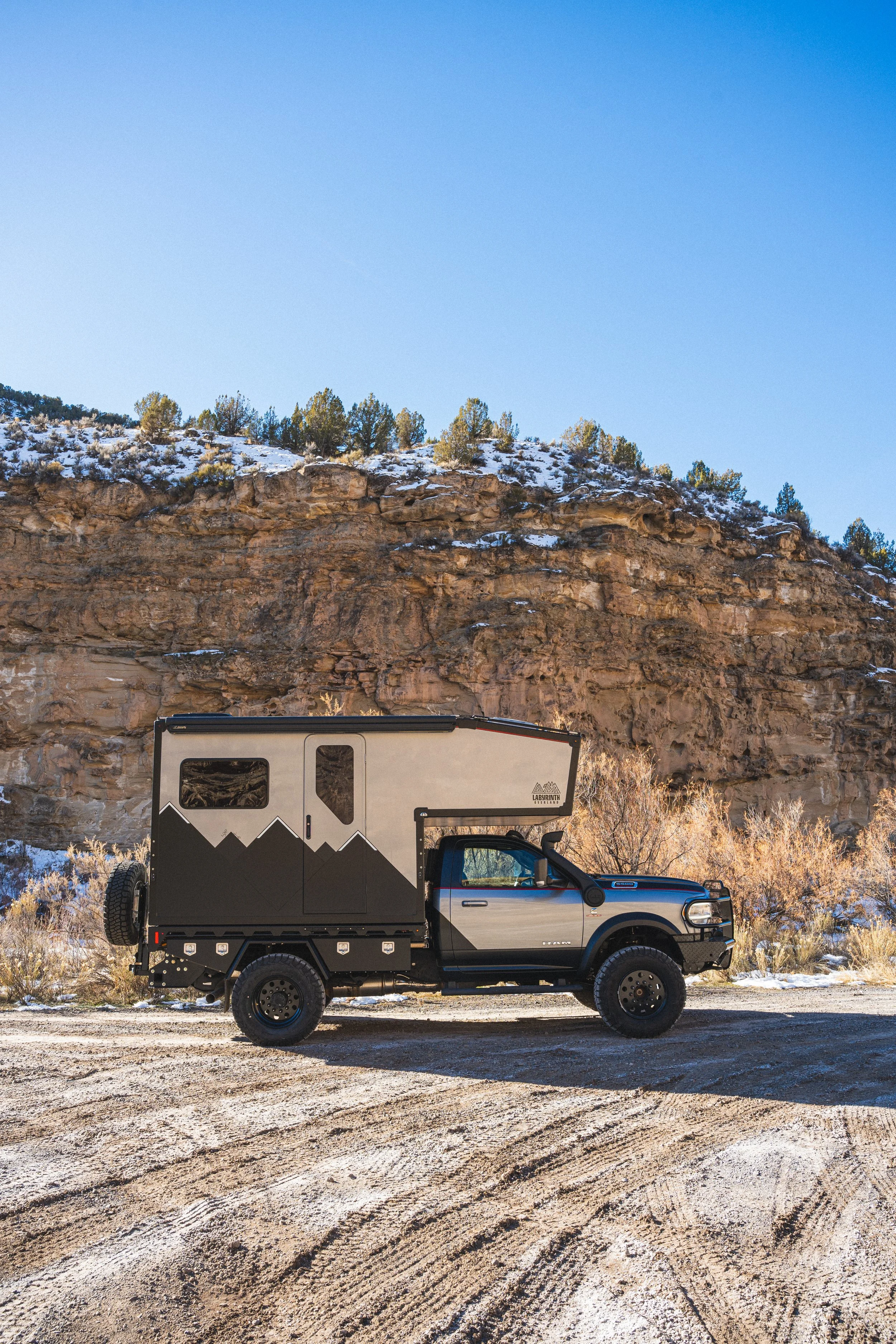A truck with an off-road camper parked on a dirt road against a rocky landscape with a cliff and sparse trees, under a clear blue sky.