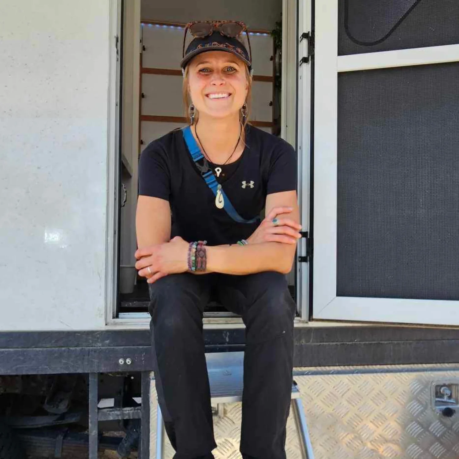 A smiling woman sitting on the steps outside a camper or RV, wearing black clothing, a baseball cap, sunglasses on her head, jewelry, and a lanyard with a shell pendant.