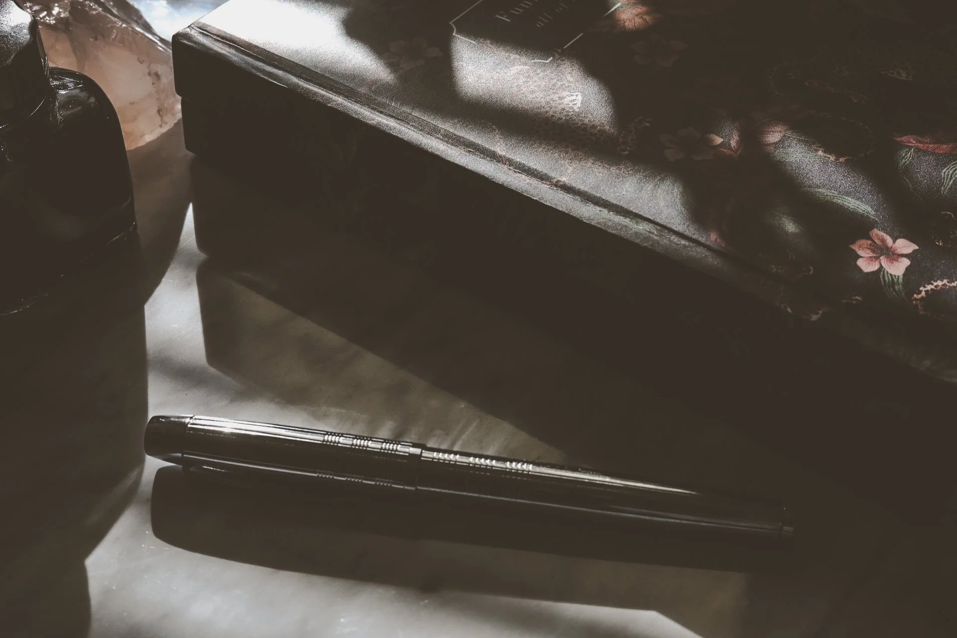 A black pen, a black Papier journal with floral patterns, and dark ink bottles are arranged on a reflective marble surface.