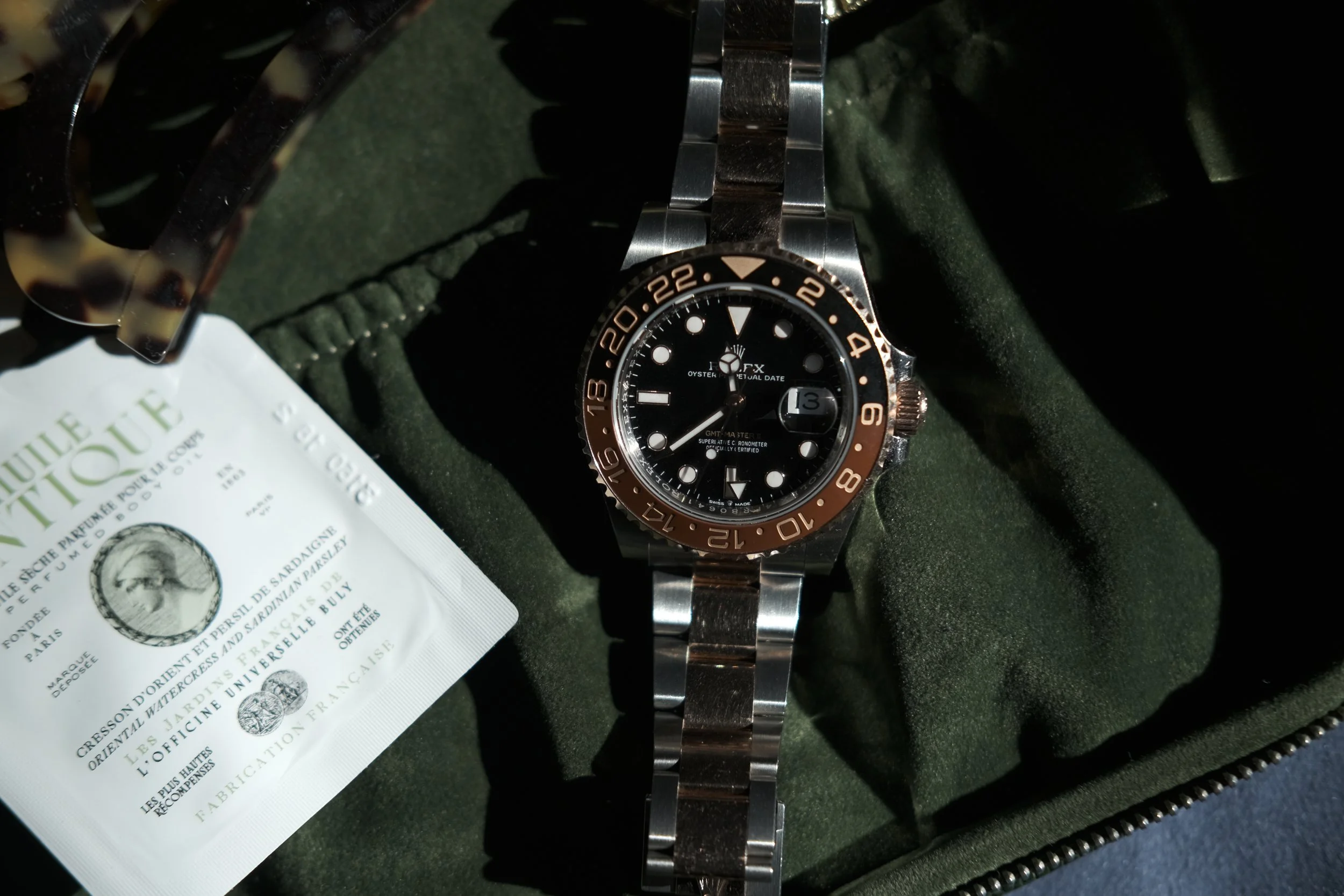 A luxury wristwatch with a black face and a two-tone bezel resting inside a green case. The watch has a metal band and displays the date. Part of a white packet with French text is visible nearby.