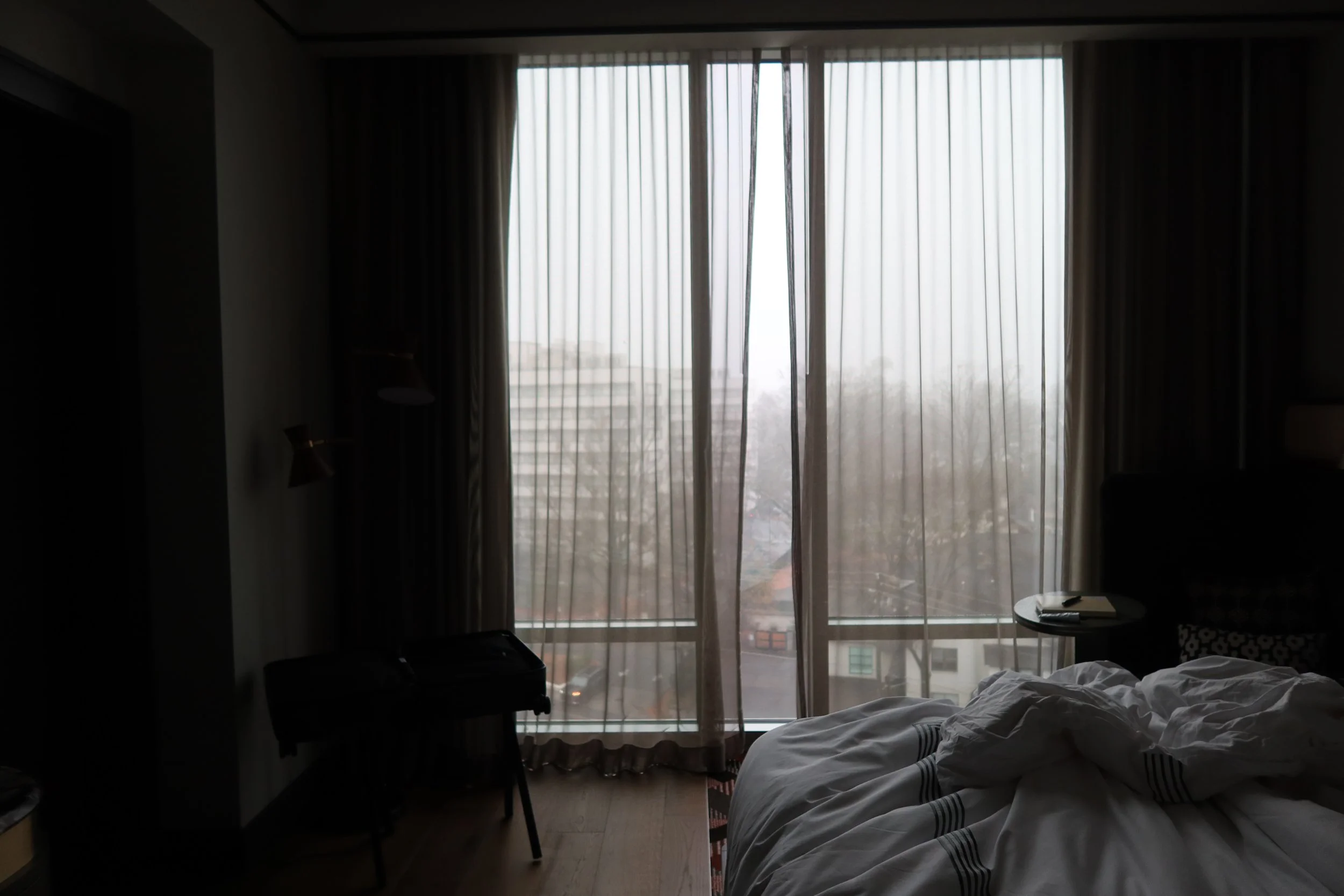 Darkened hotel room with a large window covered by sheer curtains, a disheveled bed in the foreground, a small round side table with a notebook and pen, and a chair with a suitcase underneath.