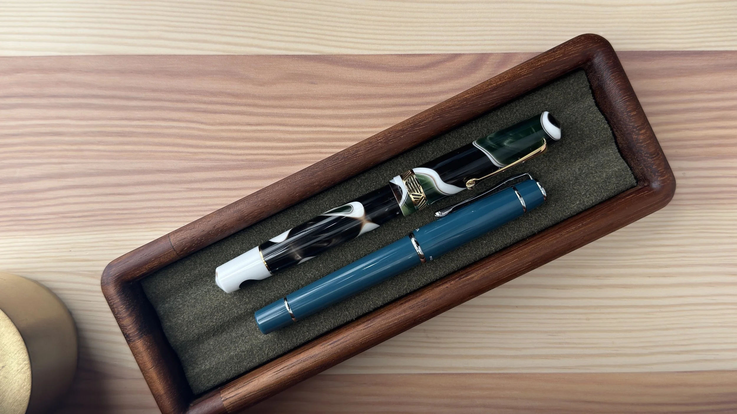 Two fountain pens, one with a marbled black and white design and the other in solid teal, placed in a wooden pen case on a light wood surface.