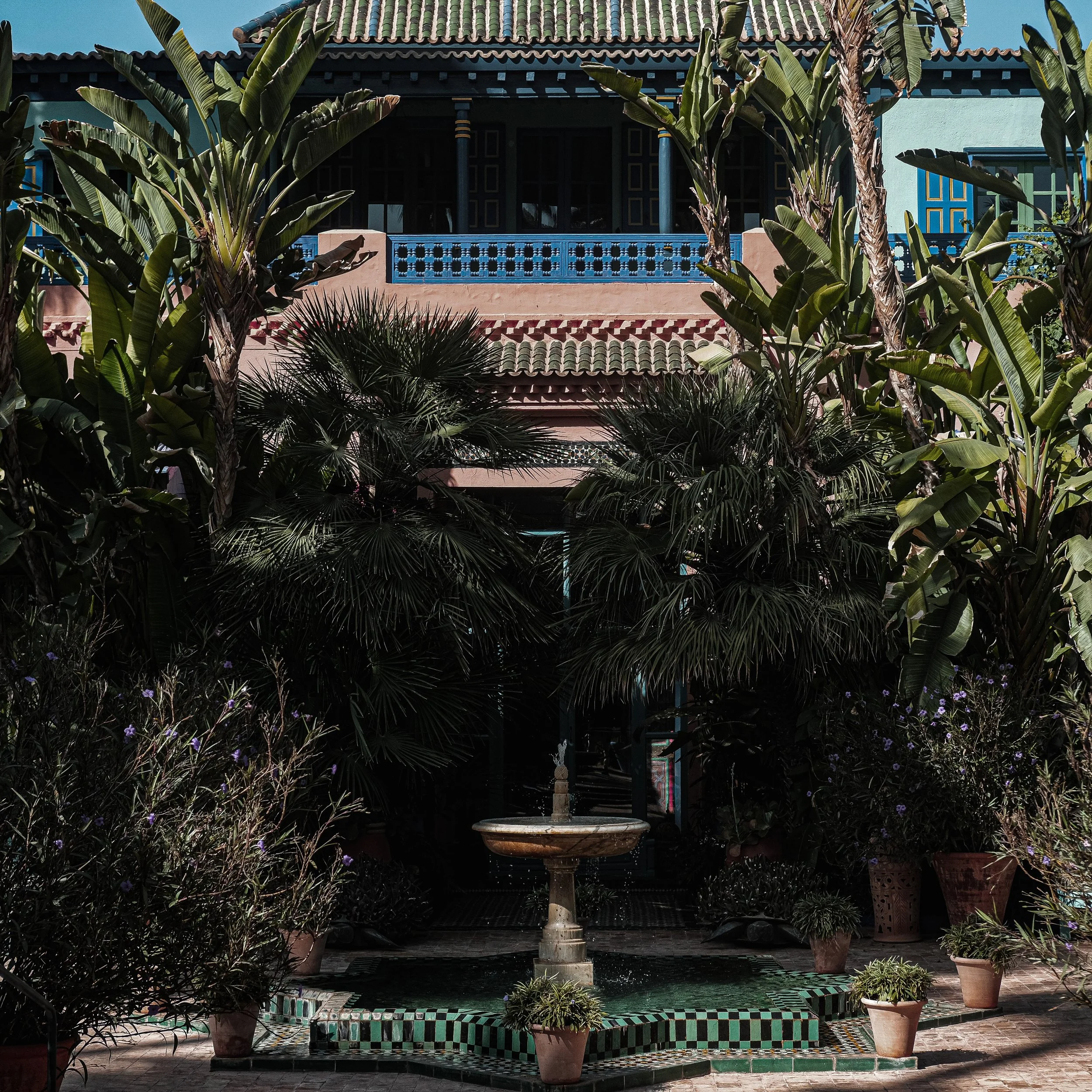 A lush courtyard with tropical plants surrounding a small fountain, underneath a building with Spanish-style architecture, including a green-tiled roof and blue accents.
