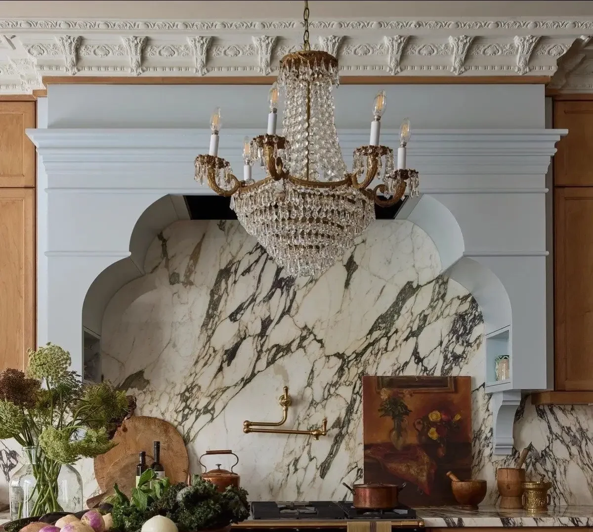 Elegant kitchen with a crystal chandelier hanging above a marble backsplash and counter. Decorative items include a painting, bowls, and kitchen utensils, with a variety of fresh vegetables and herbs on the counter.