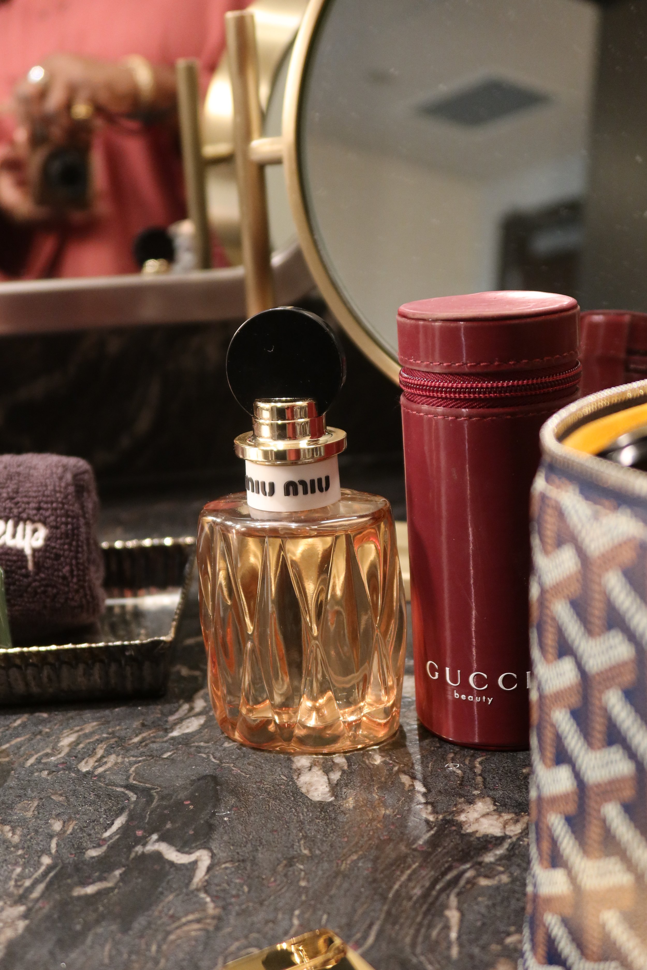 A perfume bottle with a black circular cap and a white band labeled 'Miu Miu' on a black marble countertop, alongside a red Gucci beauty container, a patterned bag, and a mirror reflecting the scene.
