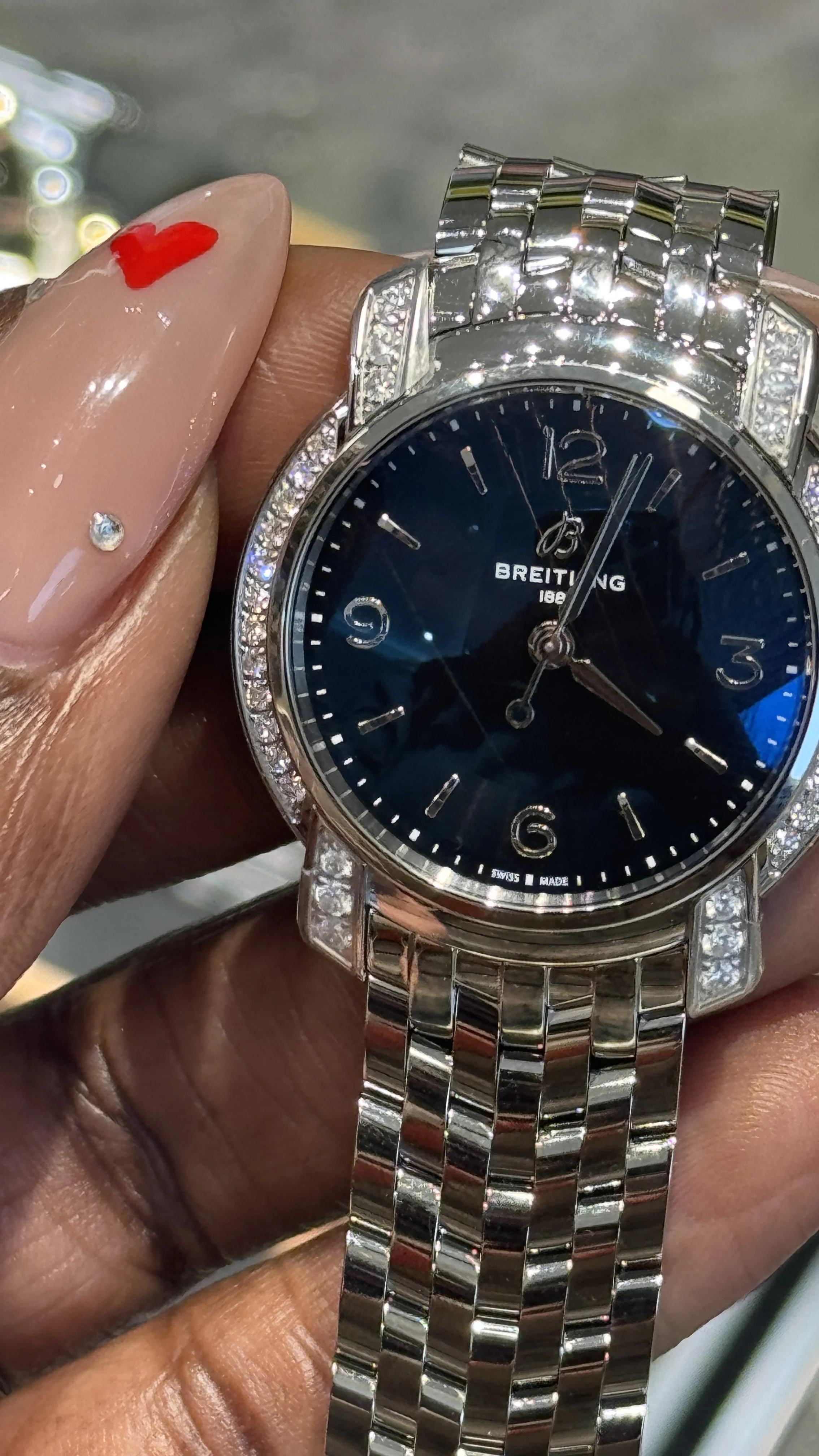 A person's hand holding a silver Breitling wristwatch with a dark blue face and diamond accents around the bezel and on the band.