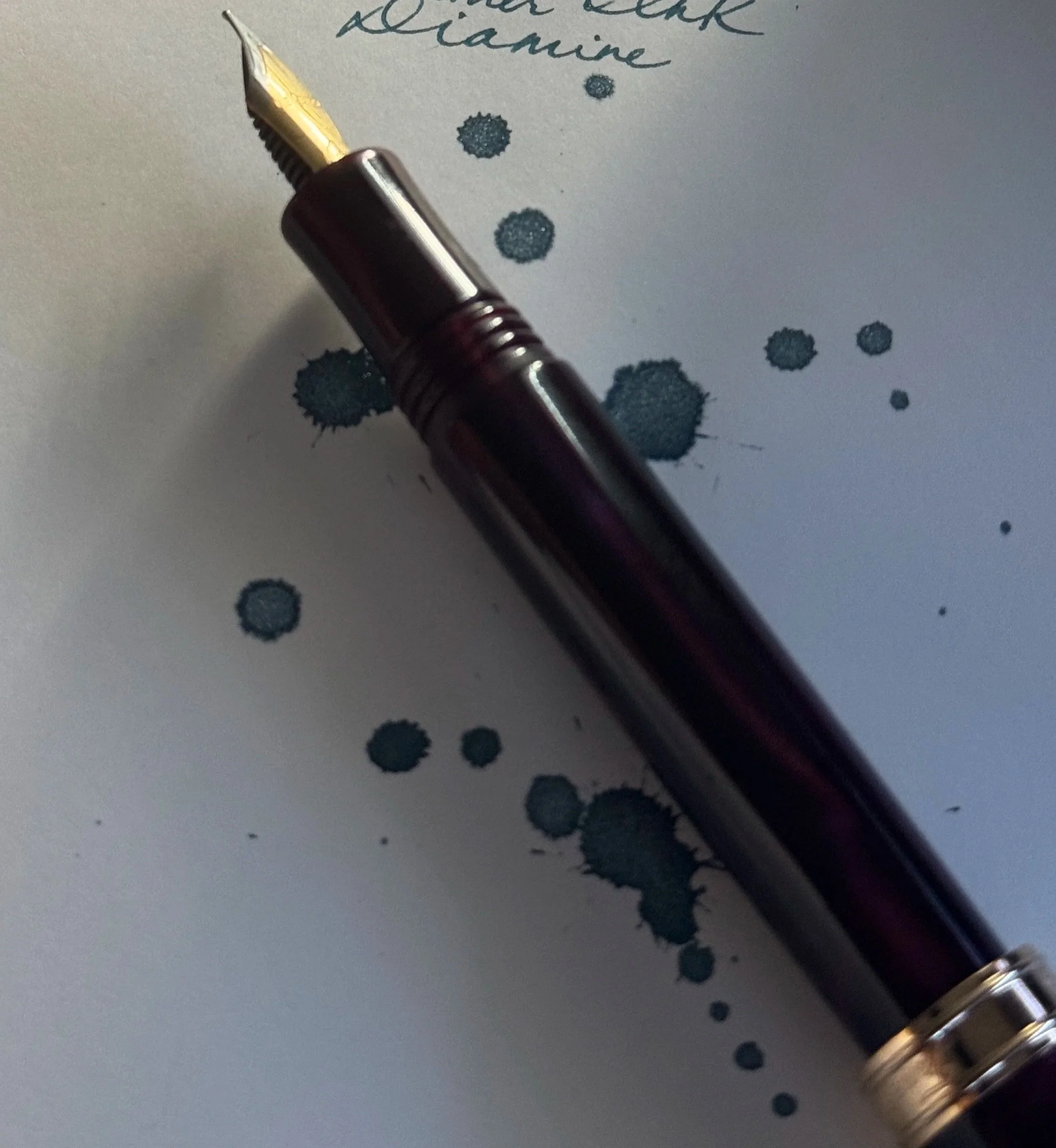 Close-up of a fountain pen resting on a sheet of paper with blue ink splatters and handwritten text that reads 'Writer’s Blood Diamine.’