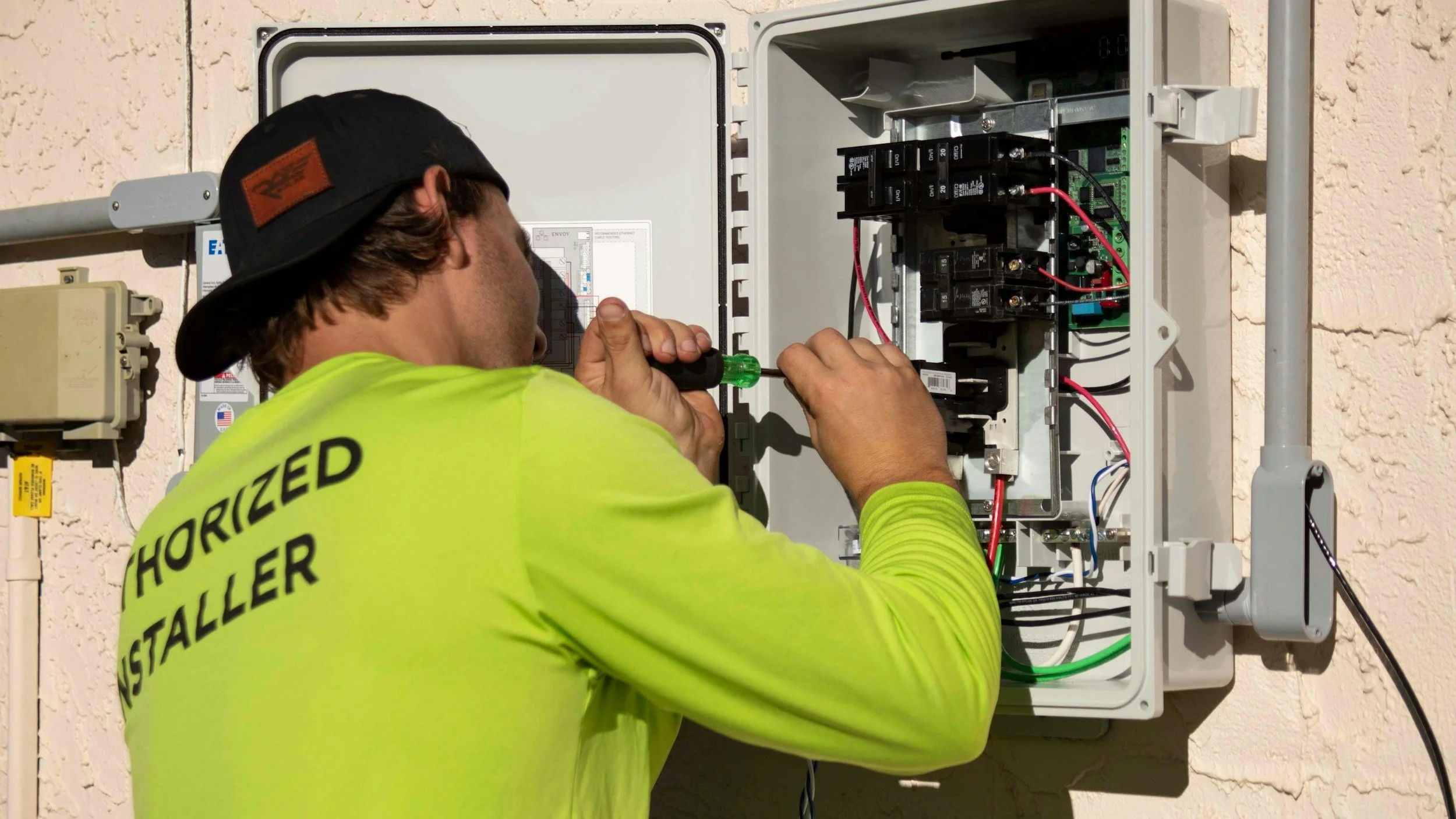 A technician wearing a bright yellow shirt and black cap inspecting an electrical box with a screwdriver.