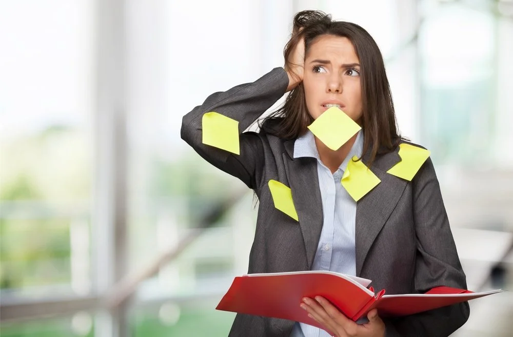 A brown-haired woman with a gray vest and light blue shirt looking anxious with post-it notes all over her chest, and one in her mouth