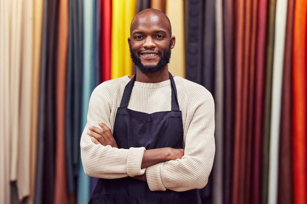 A man in a black apron and white shirt stands with folded arms looking straight ahead on a yellow background
