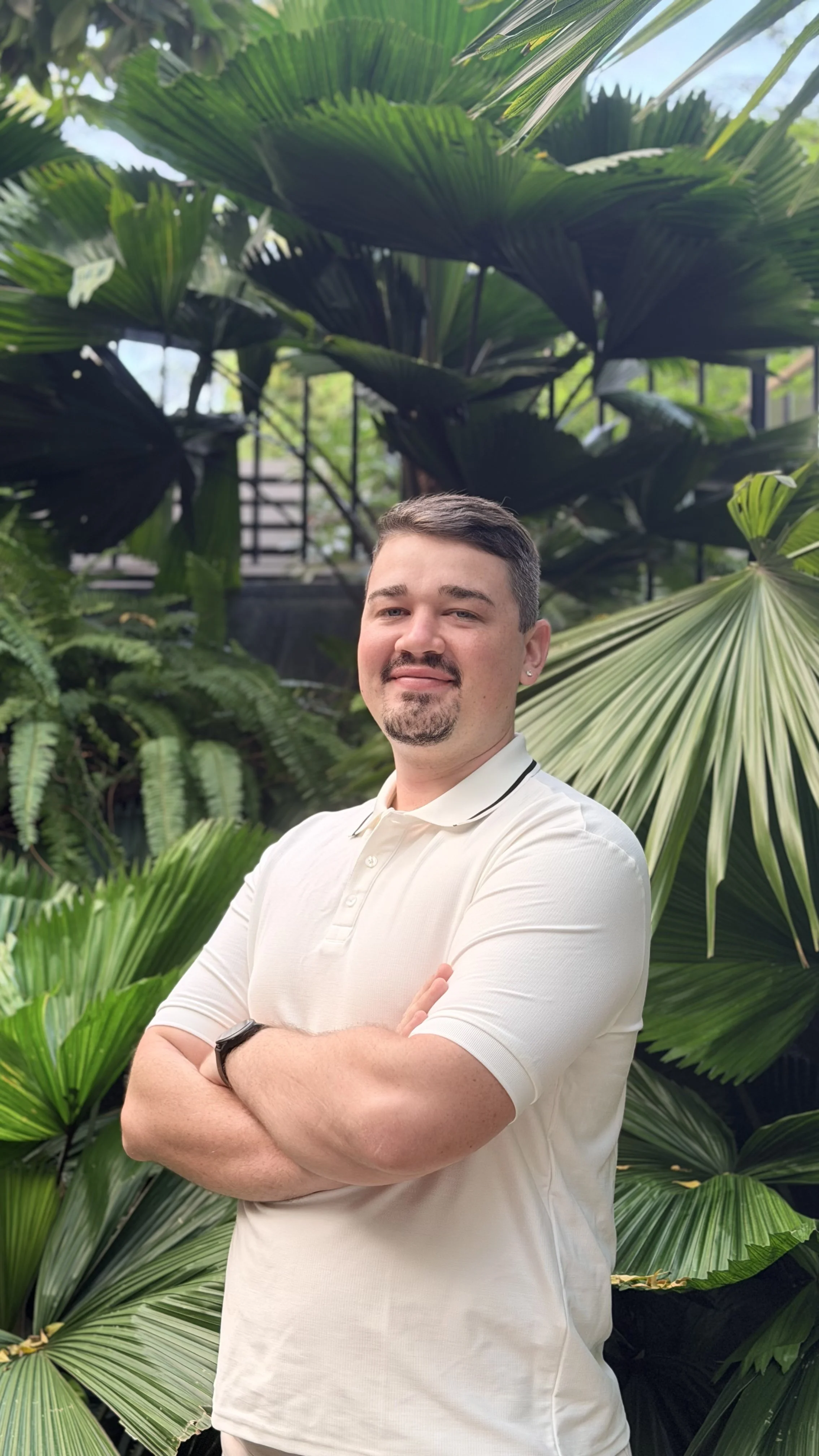 Man with crossed arms standing in front of lush green tropical plants.