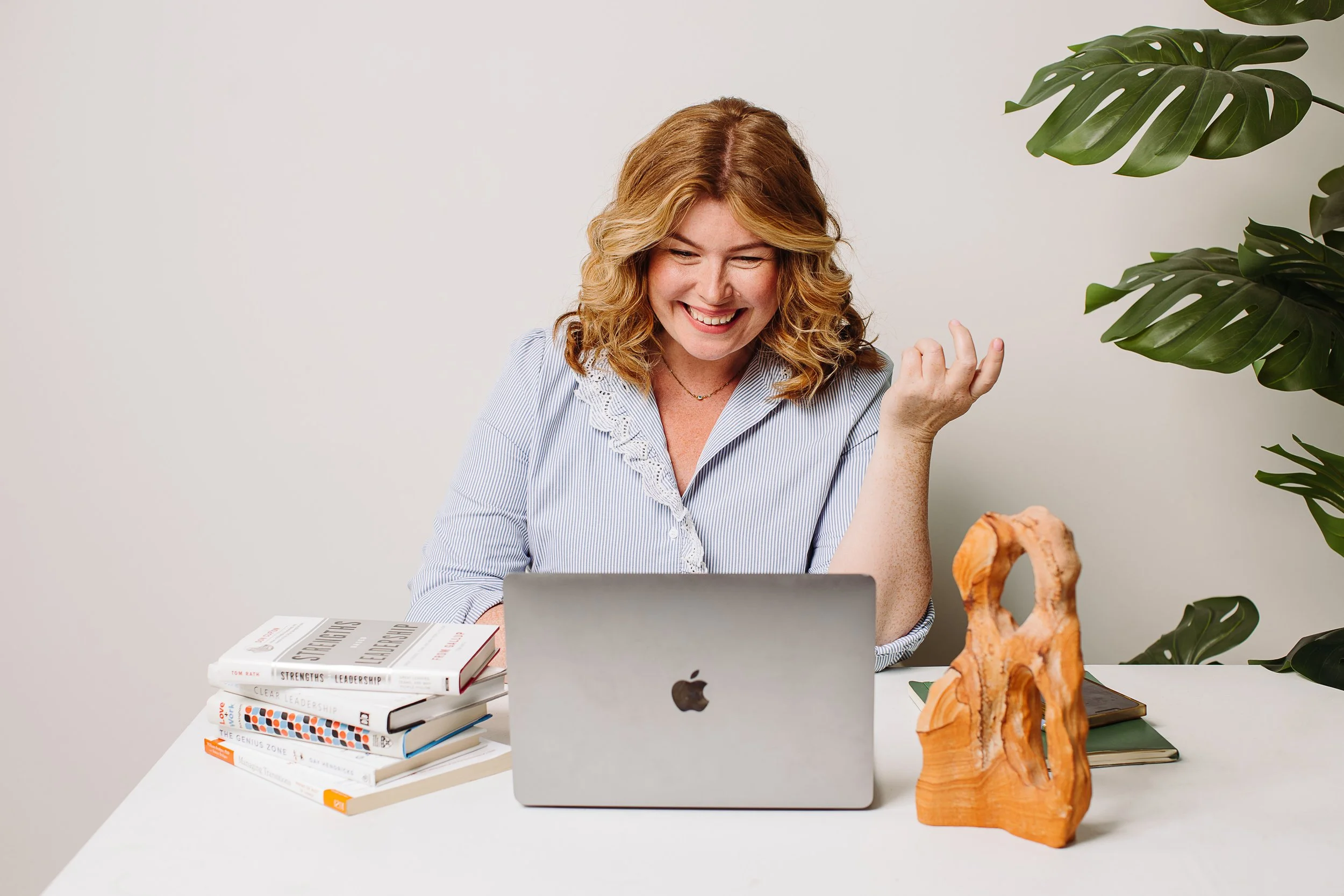 A woman with red hair and a striped shirt smiling and looking at her laptop in a bright, minimalistic office space, with a stack of books, a wooden sculpture, green plants, and a notebook on her desk.