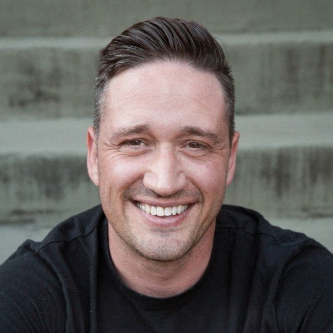 A smiling man with short hair in a black shirt is taking a selfie outdoors against a concrete staircase background.
