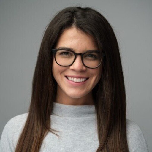 A young woman with long brown hair, wearing glasses and a light gray top, smiling against a plain gray background.