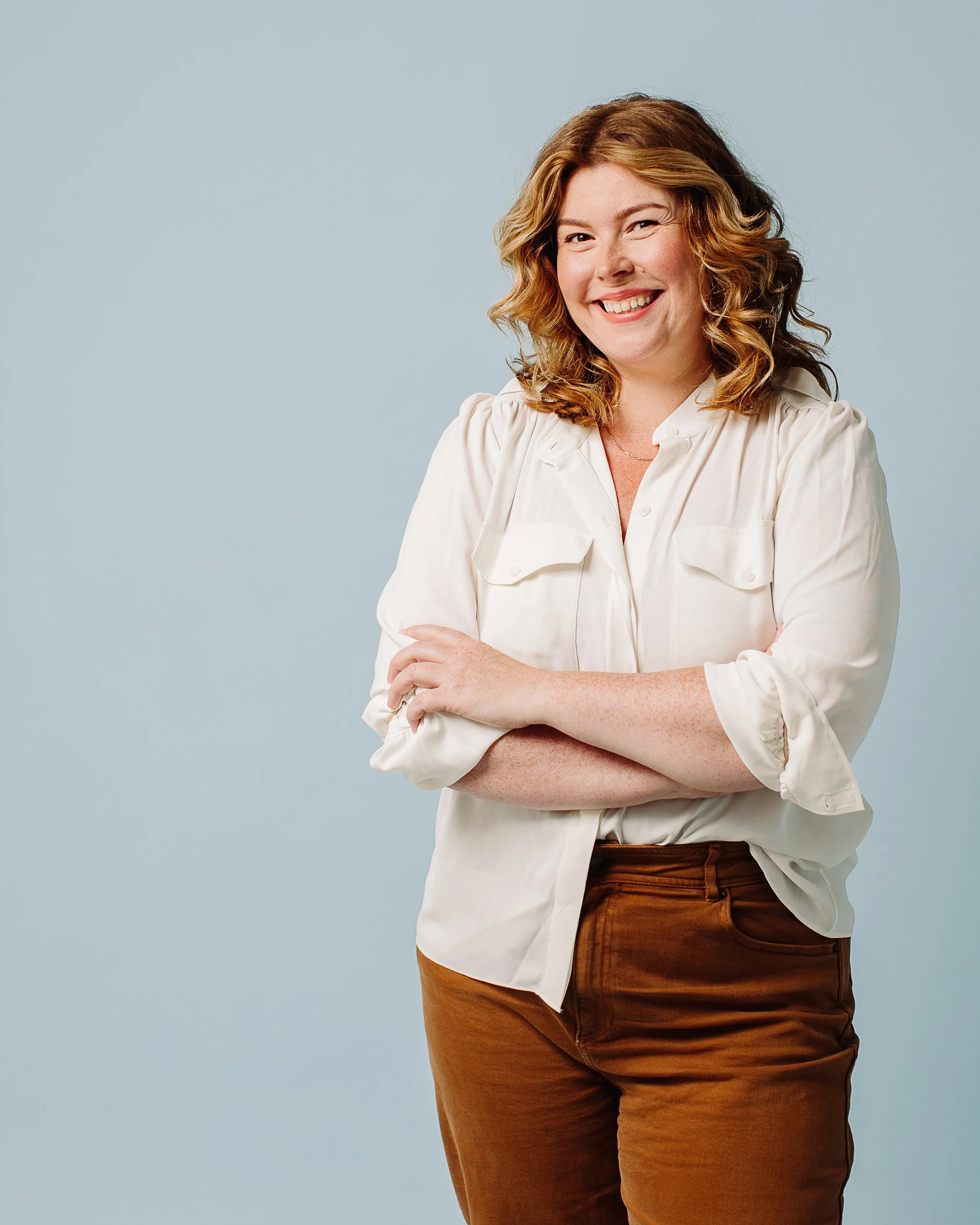 A woman with curly, shoulder-length hair smiling at the camera, wearing a cream-colored button-up shirt and brown pants, standing against a light blue background.