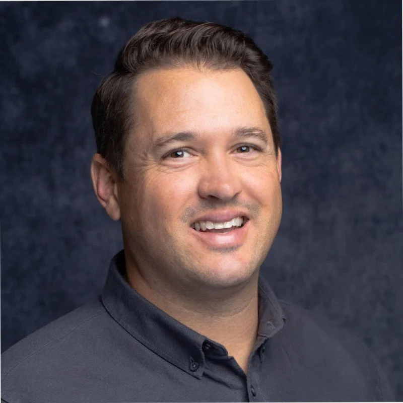 Headshot of a smiling man with short dark hair, wearing a dark button-up shirt, against a dark textured background.