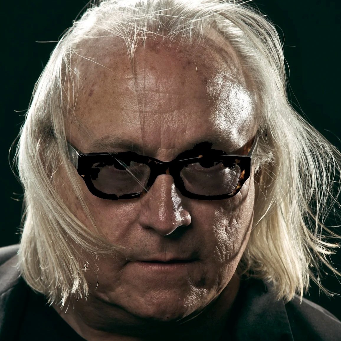 Close-up of an elderly man with long white hair, wearing dark glasses, with a serious expression and a dark background.