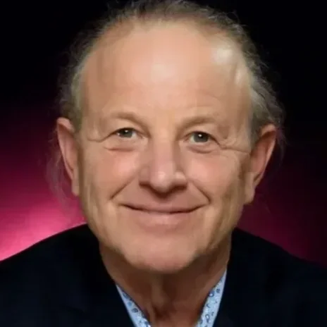Close-up of a smiling older man with gray hair, wearing a dark blazer and a patterned blue shirt, against a dark and pinkish background.