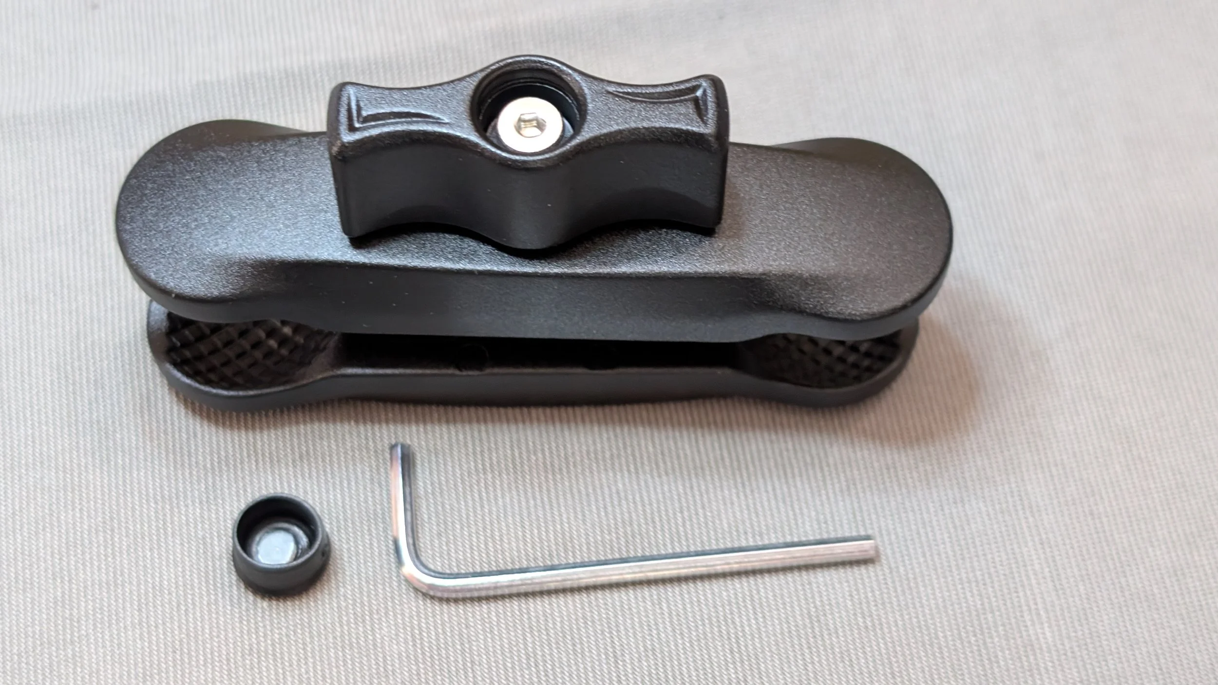 A black clamp with a textured handle, a small black rubber or plastic component, a small metal L-shape tool, and a tiny screw or bolt on a light-colored surface.