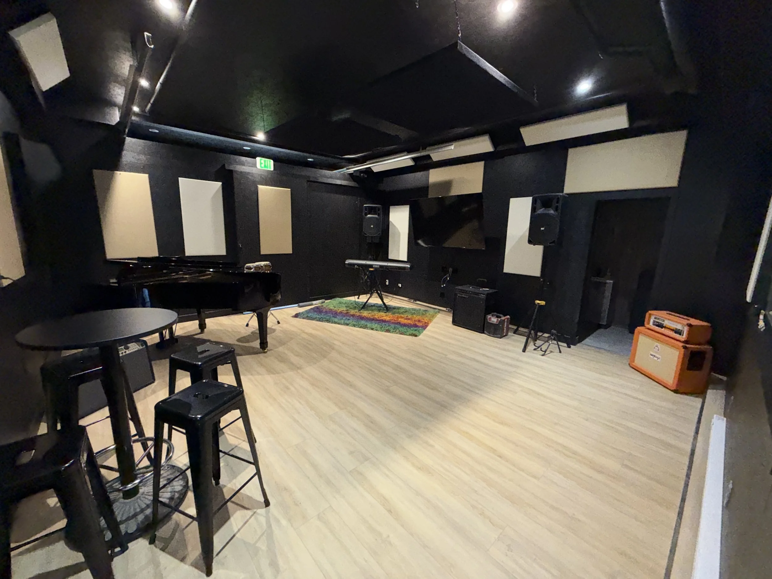 Empty music rehearsal room with musical instruments and equipment, including a grand piano, a keyboard on a stand, speakers, guitar amplifiers, and a small table with four chairs. The room has black walls with acoustic panels and a wooden floor.