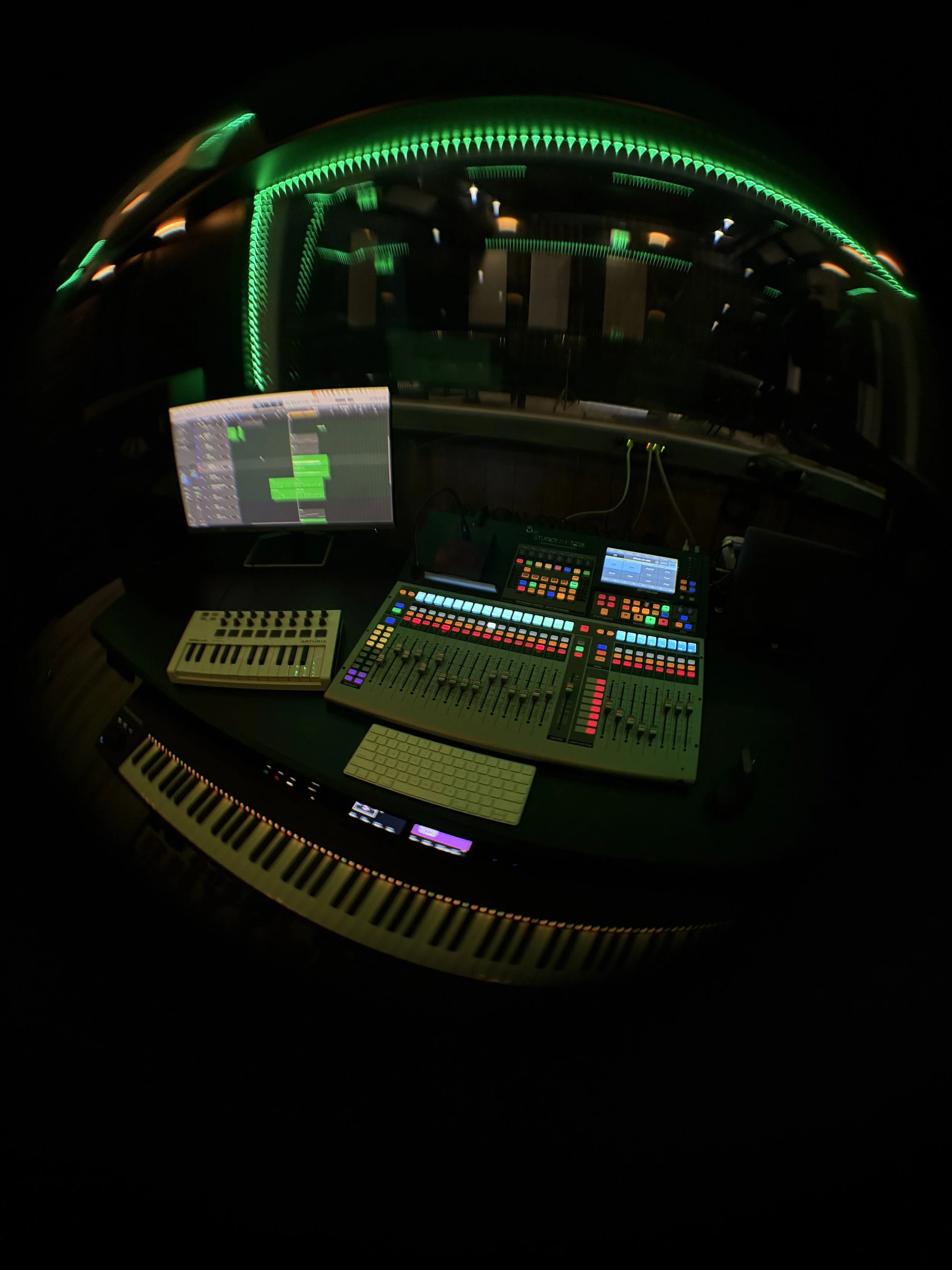 Music production studio setup with a digital mixing console, a computer monitor showing a digital audio workstation, a small MIDI keyboard, and other electronic music equipment, illuminated with green LED lights.