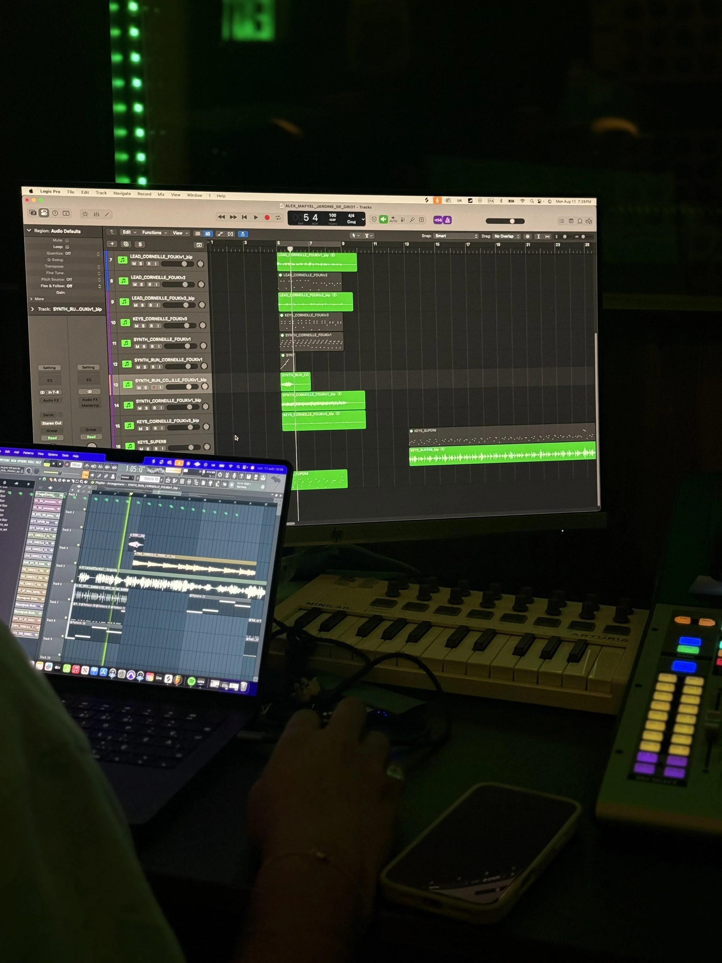 A computer setup with two screens displaying music production software, a MIDI keyboard, a smartphone, and a person's hand adjusting controls in a dark environment.