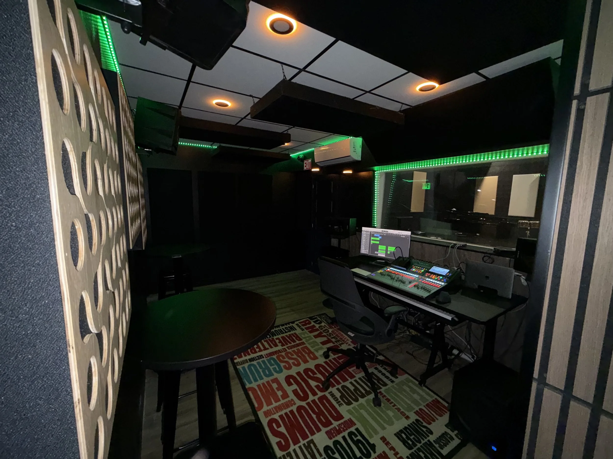Recording studio control room with mixing console, computer monitors, black chair, black and beige patterned carpet, and green LED lighting accents.