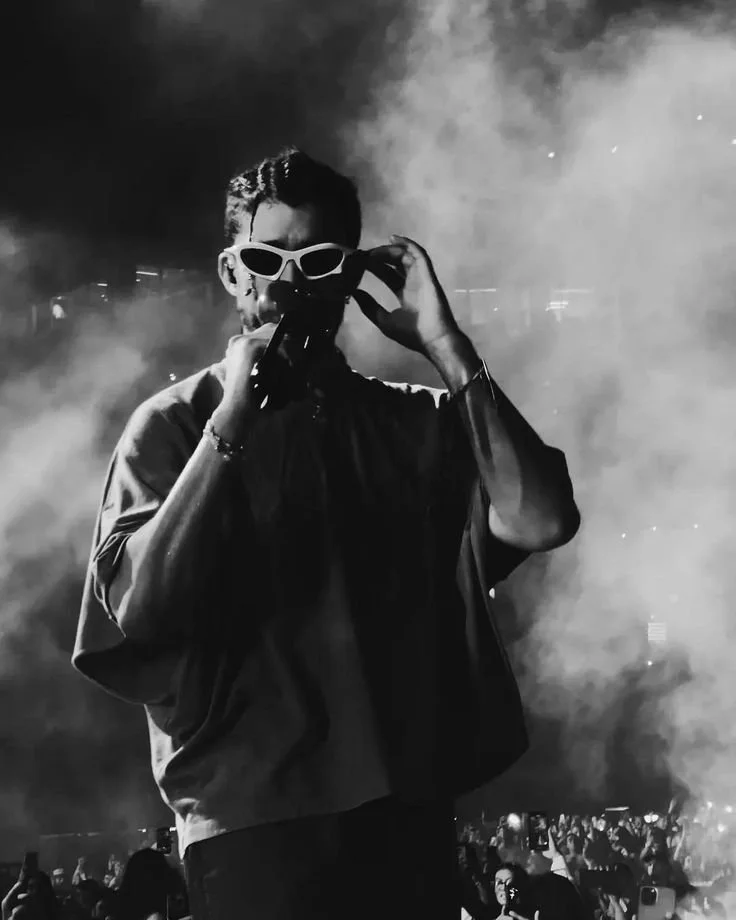 A person with short hair wearing white sunglasses, holding a microphone, in a smoky environment with an audience in the background.