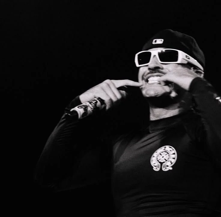 A man wearing a black baseball cap, white sunglasses, and a black shirt with a logo, smiling and adjusting his headphones.