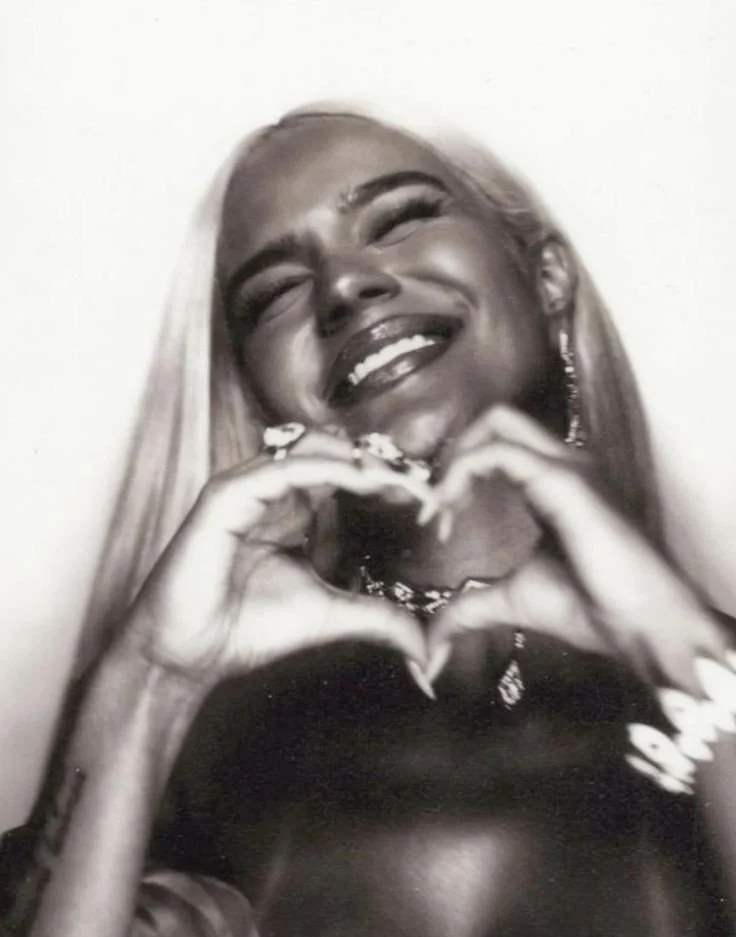 A woman with long blonde hair smiling and making a heart shape with her hands.