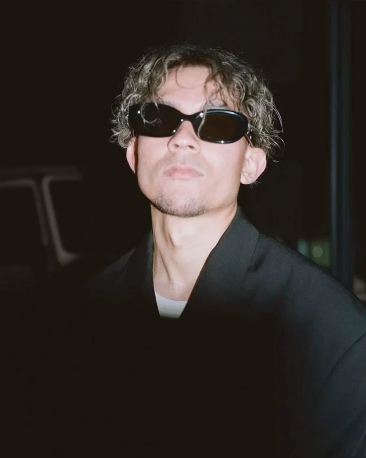 A man with curly hair wearing dark sunglasses and a black jacket, standing in a dimly lit setting.