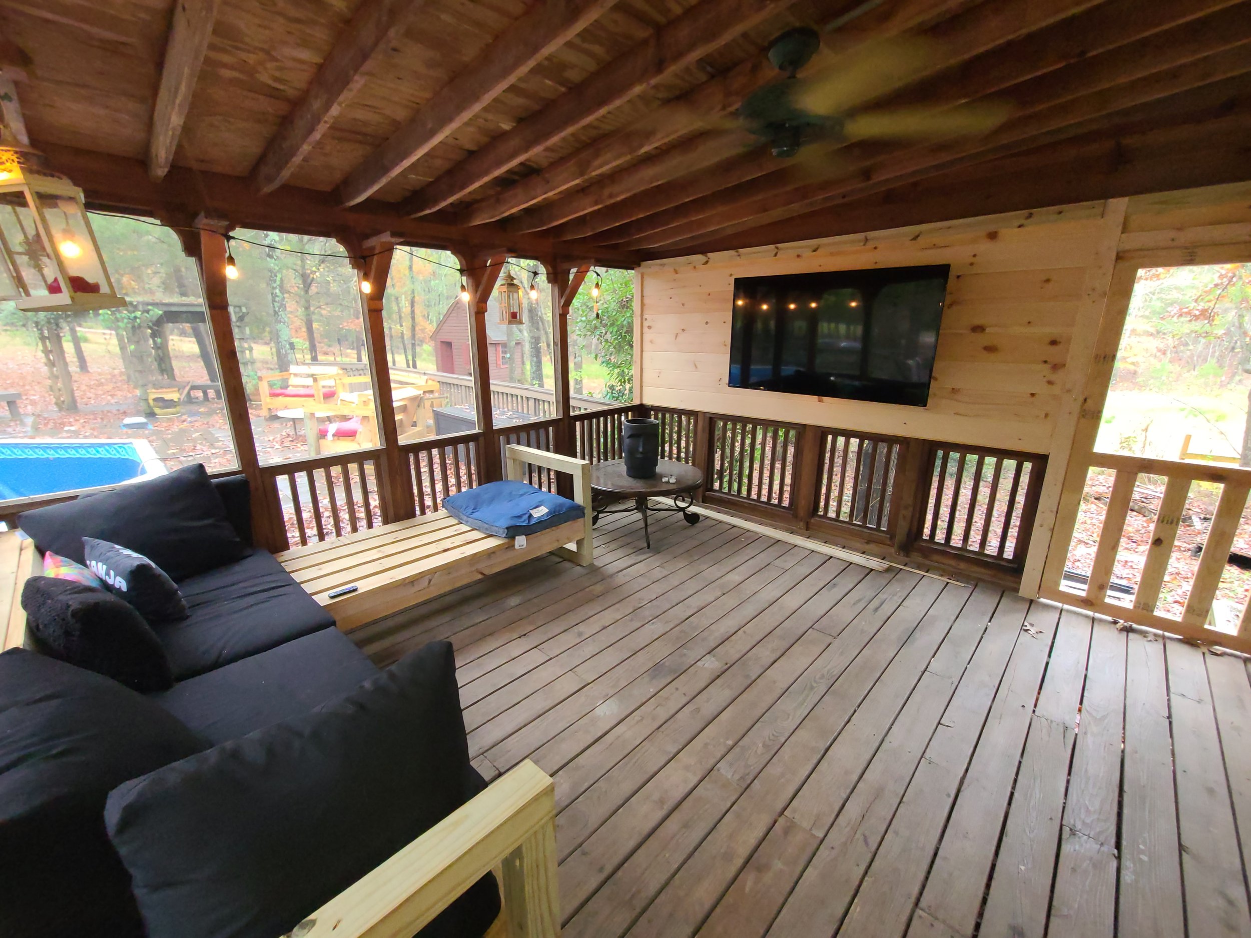 A screened porch with wooden floors and ceiling, furnished with a black couch with pillows, a rustic bench with a blue cushion, a round metal table, and a large flat-screen TV mounted on the wooden-paneled wall. Outside, trees and a backyard with a glimpse of a playhouse and a blue kiddie pool are visible.