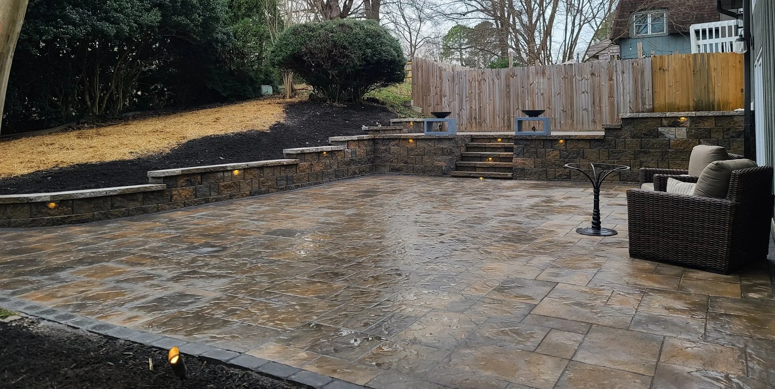 Empty outdoor patio with wet stone flooring, surrounded by a retaining wall with steps, a wooden fence, and some garden lighting. There are two chairs and a small table on the patio.