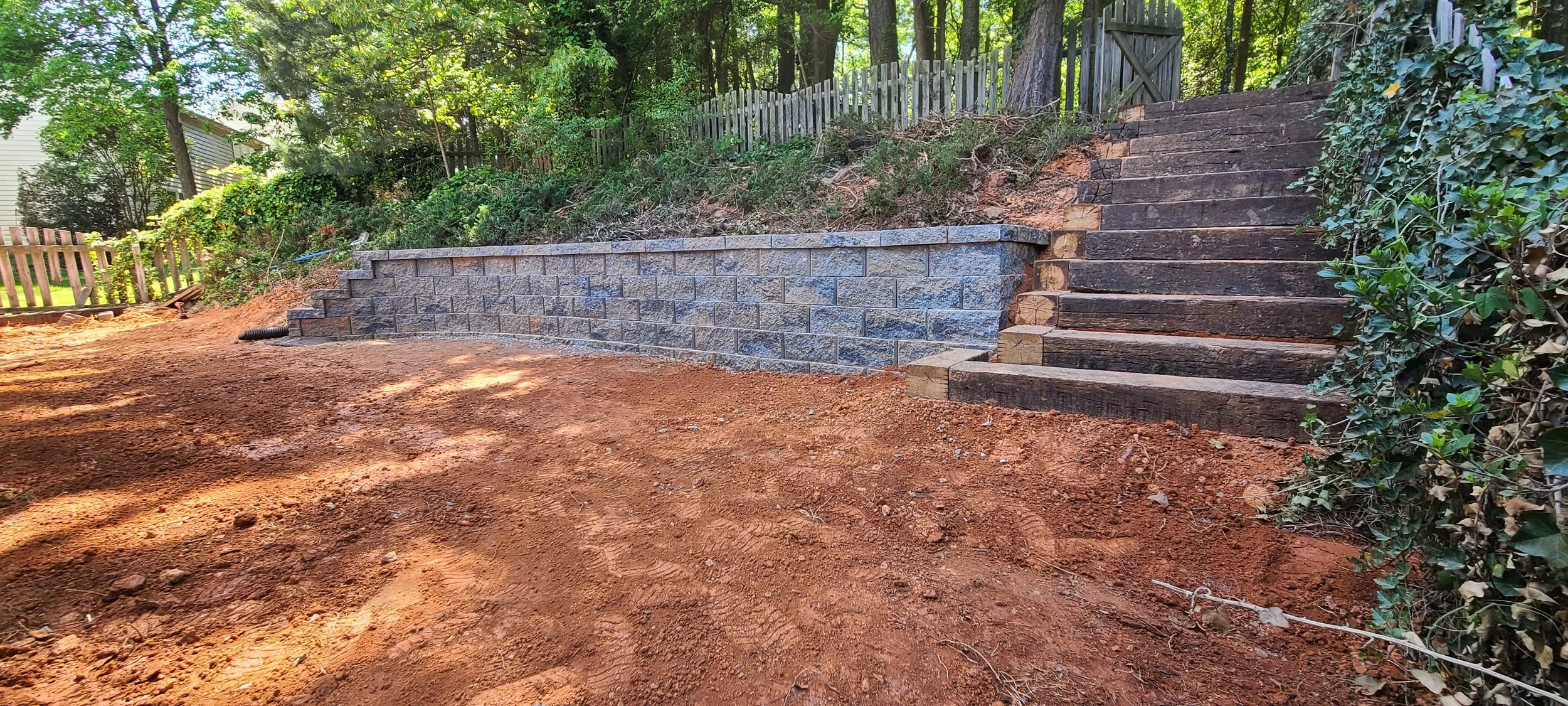 Incomplete outdoor staircase with wooden steps and a retaining wall with stone bricks, on a dirt ground, surrounded by trees and bushes.
