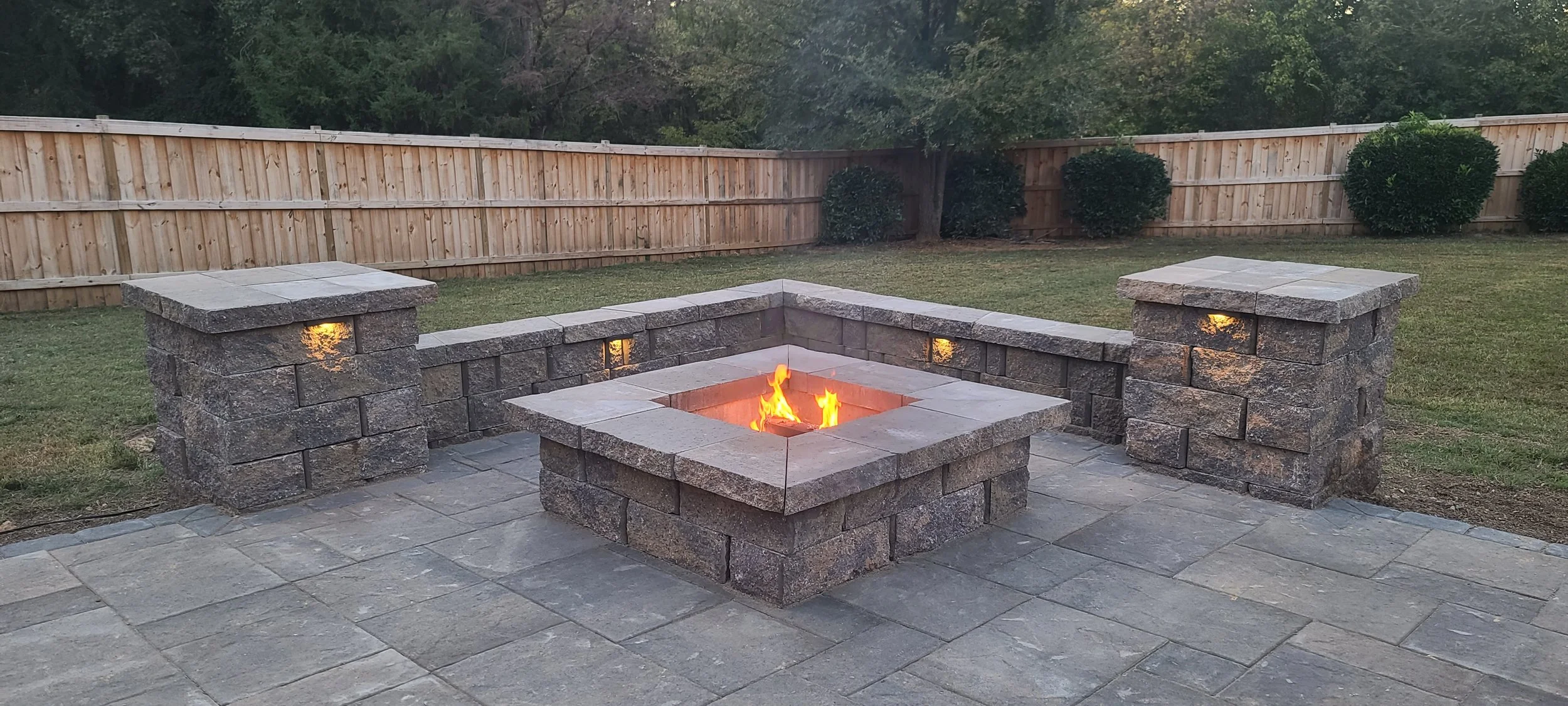 A backyard patio with a stone fire pit at center, surrounded by a low stone wall and two stone pillars. The fire is lit, and a wooden fence encloses the yard with bushes along the fence line.