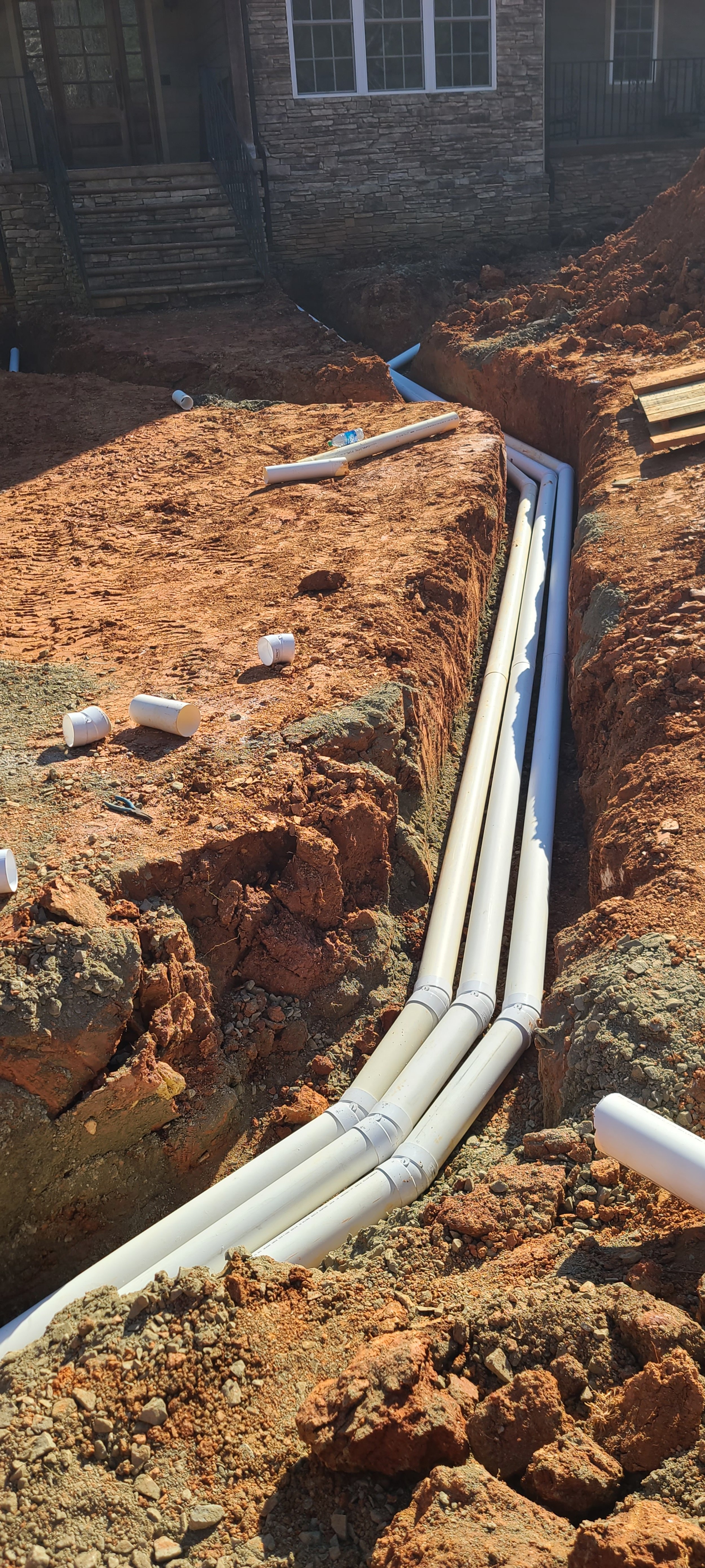 Construction site with exposed orange soil and multiple white PVC pipes being installed underground.