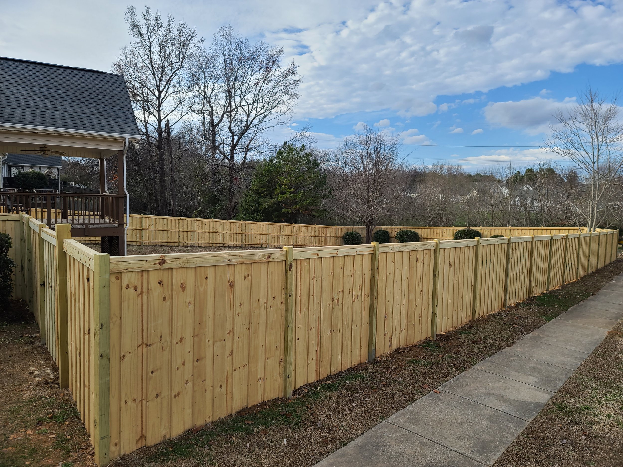 A newly installed wooden privacy fence surrounds a backyard with a concrete sidewalk on the right side. The yard has minimal grass and some bushes, with a house and porch on the left, and trees and rooftops visible in the background under a partly cloudy sky.