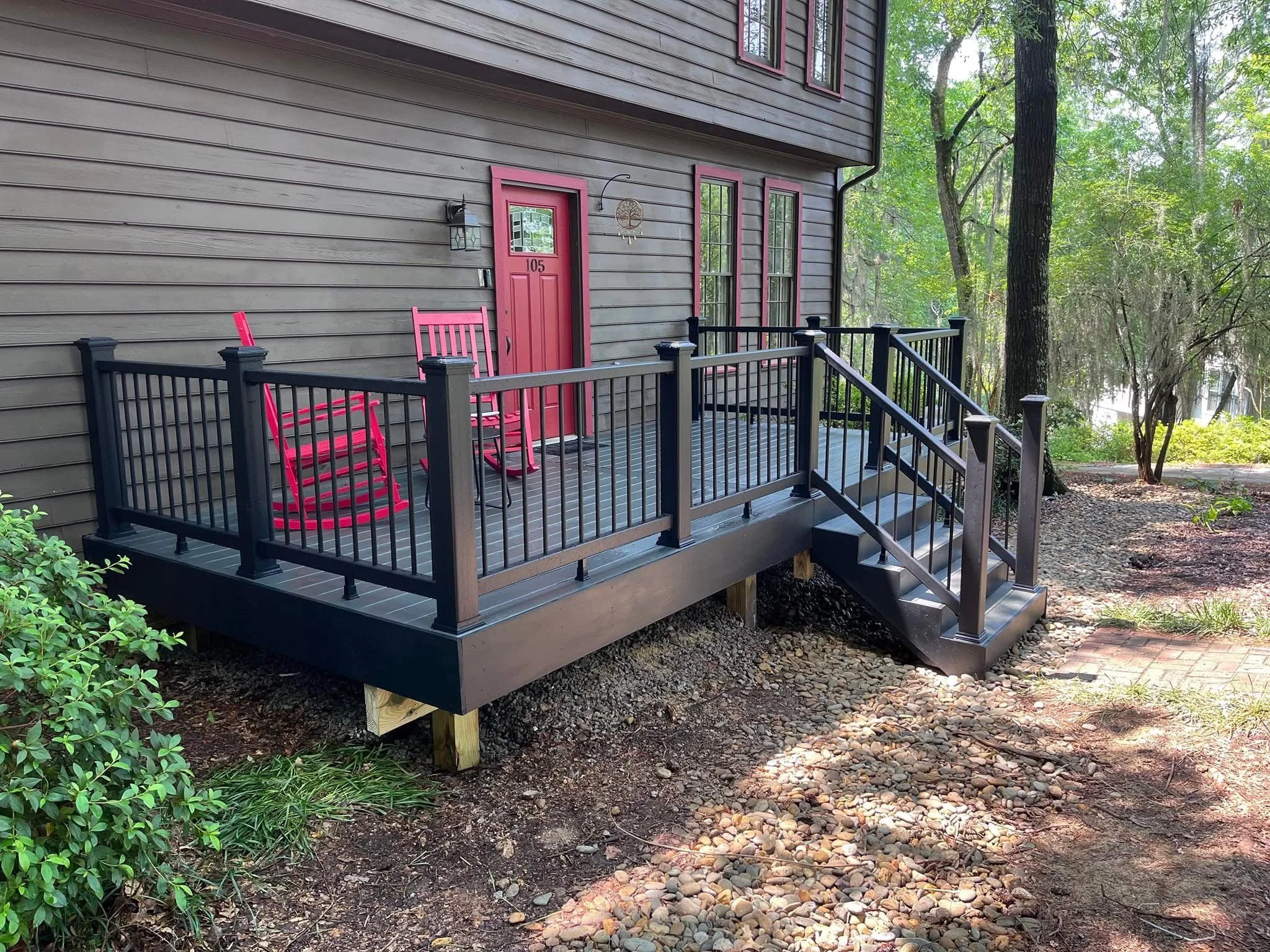 A small wooden deck attached to a house with a pink door and matching pink chairs, surrounded by trees and natural landscaping.