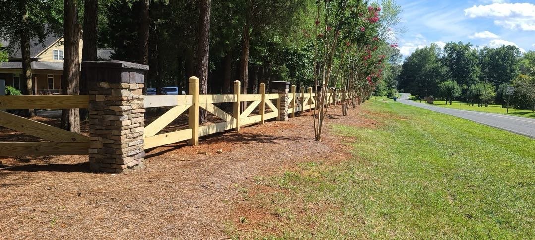 New wooden fence with stone posts along a grassy roadside with trees and houses in the background.