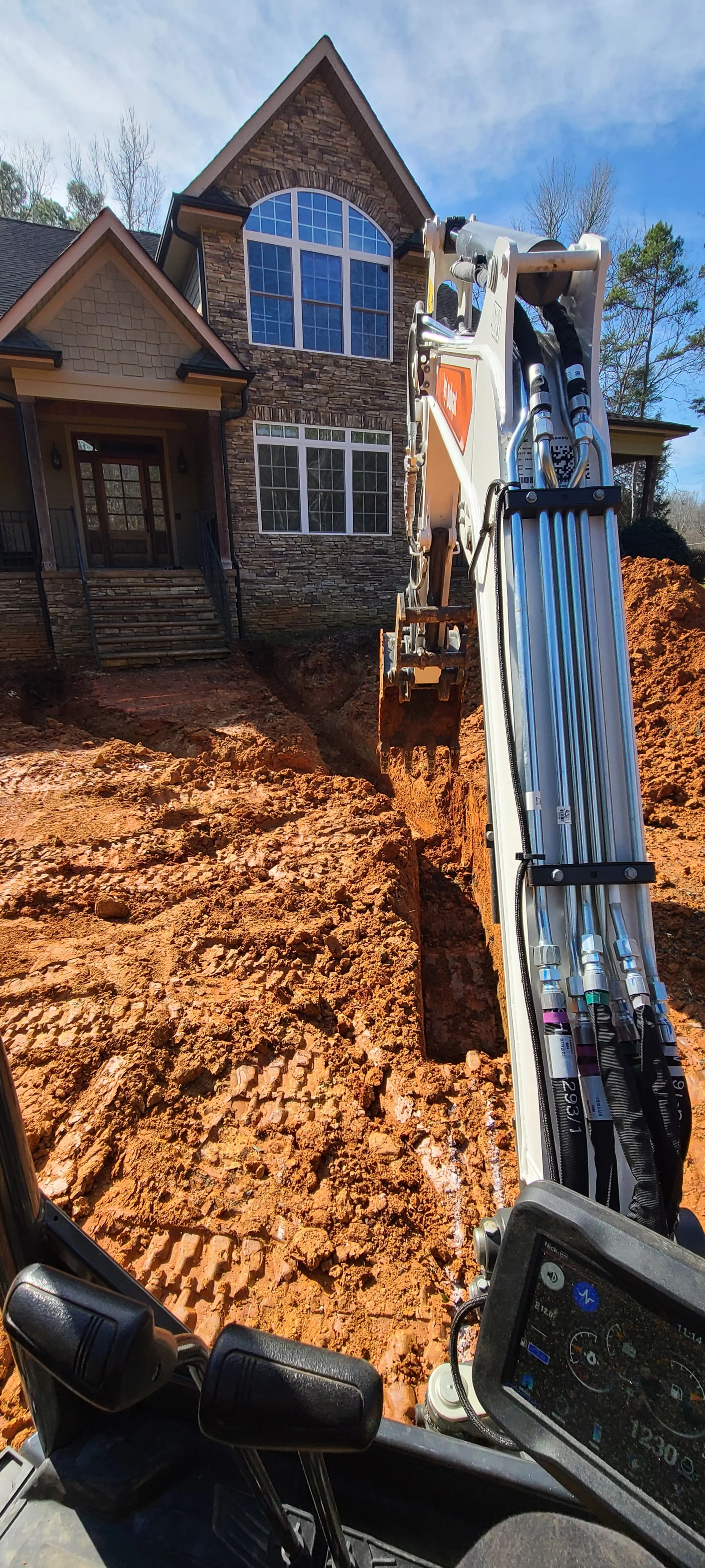 A construction excavator digging a trench in front of a two-story house with stone and siding exterior and large windows, under a partly cloudy sky.