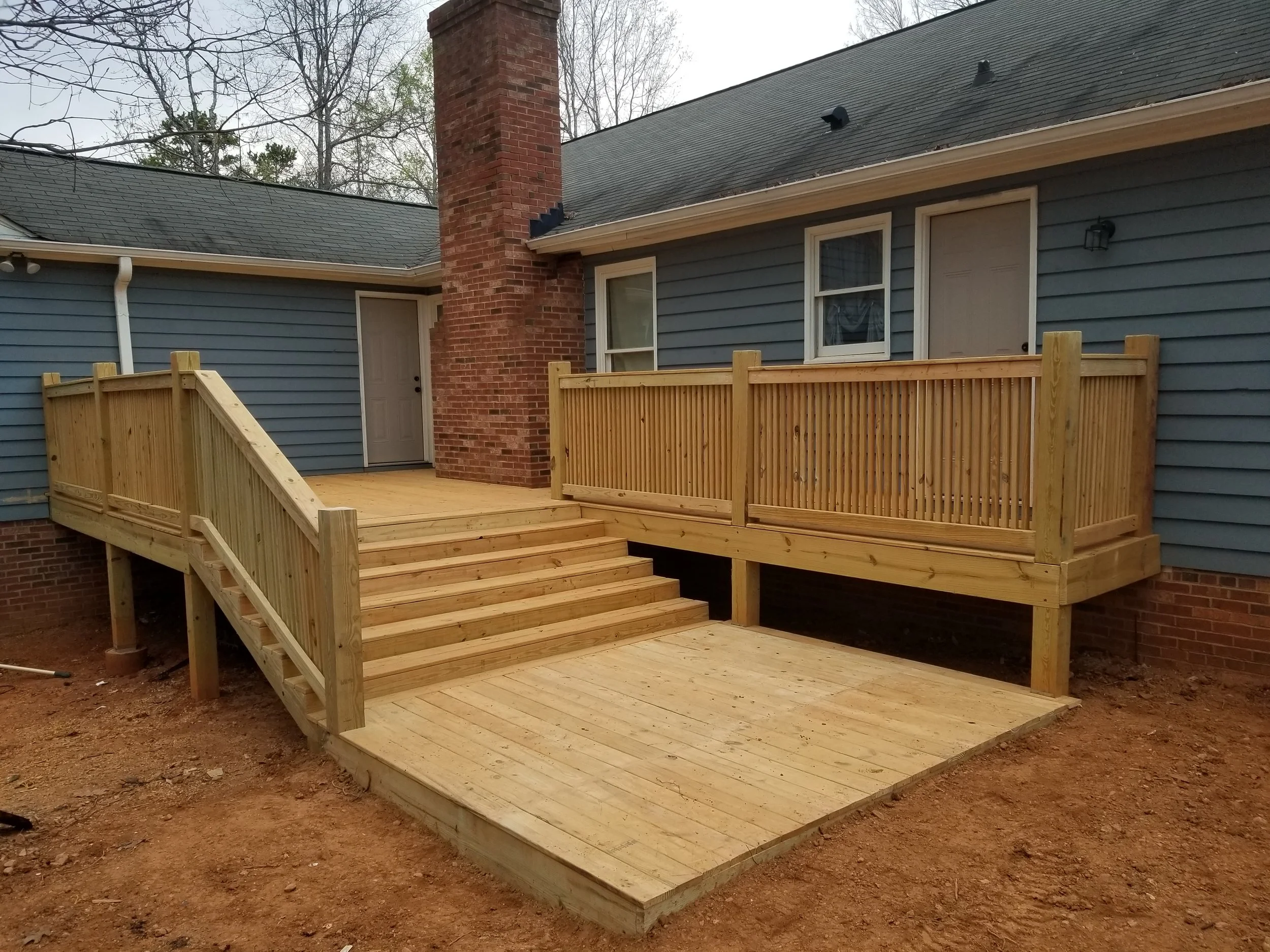 Newly built wooden deck with stairs and railing attached to a blue house with brick chimney, in a backyard, under construction.