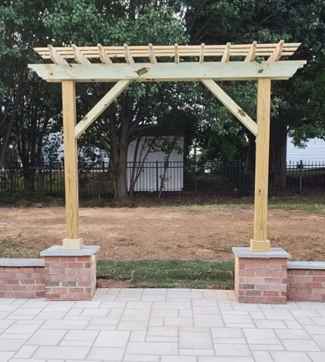 Unfinished wooden arbor with brick bases on a tiled patio, with trees and a black fence in the background.