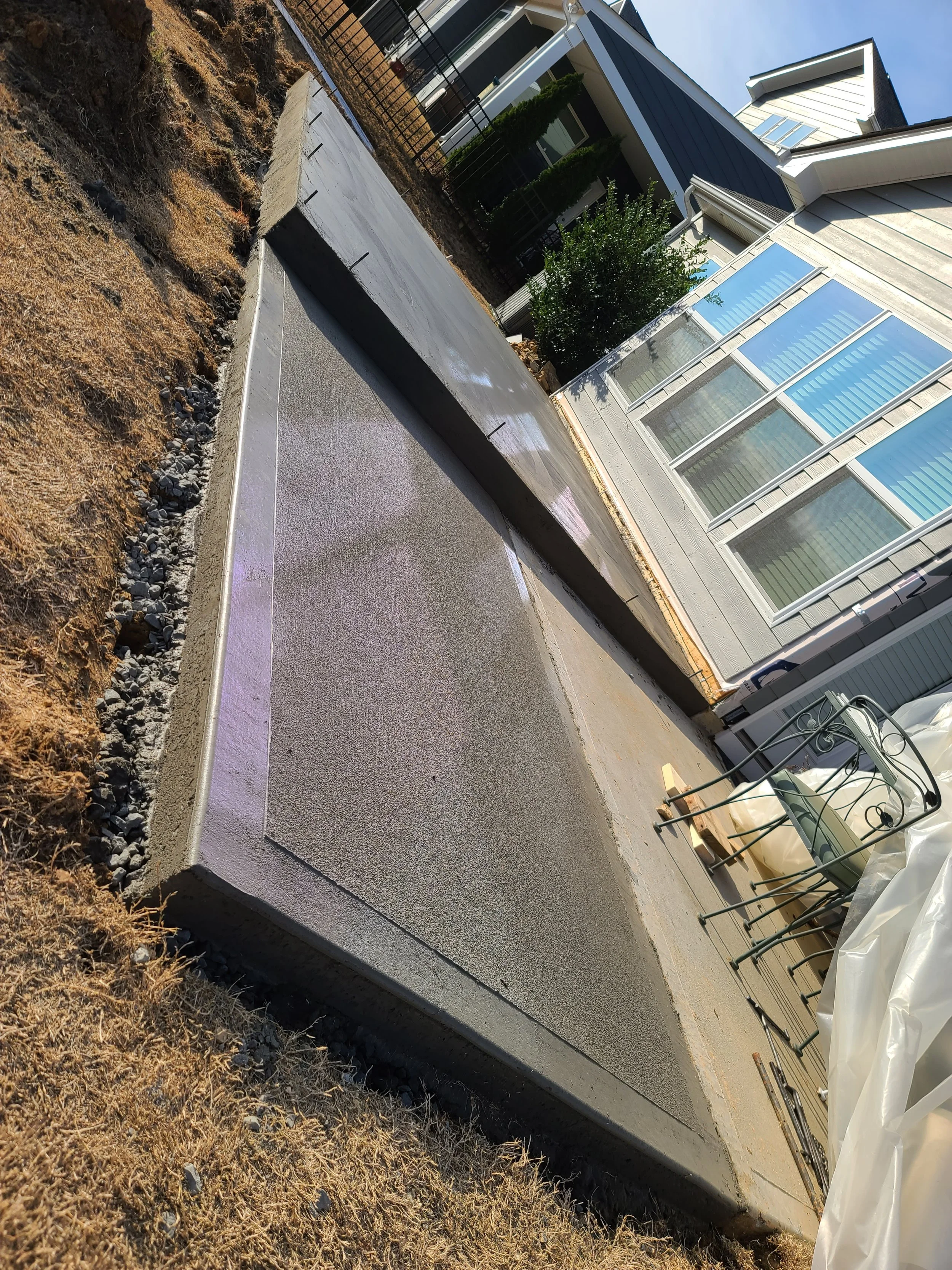 Recently poured concrete slab in a backyard, with a concrete form at one end, next to a house with large windows.