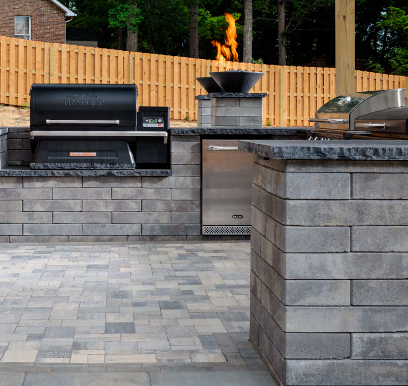 Outdoor kitchen with a black Traeger grill, a stainless steel Delsa appliance, and a fire pit on a brick paved patio with a wooden fence and trees in the background.