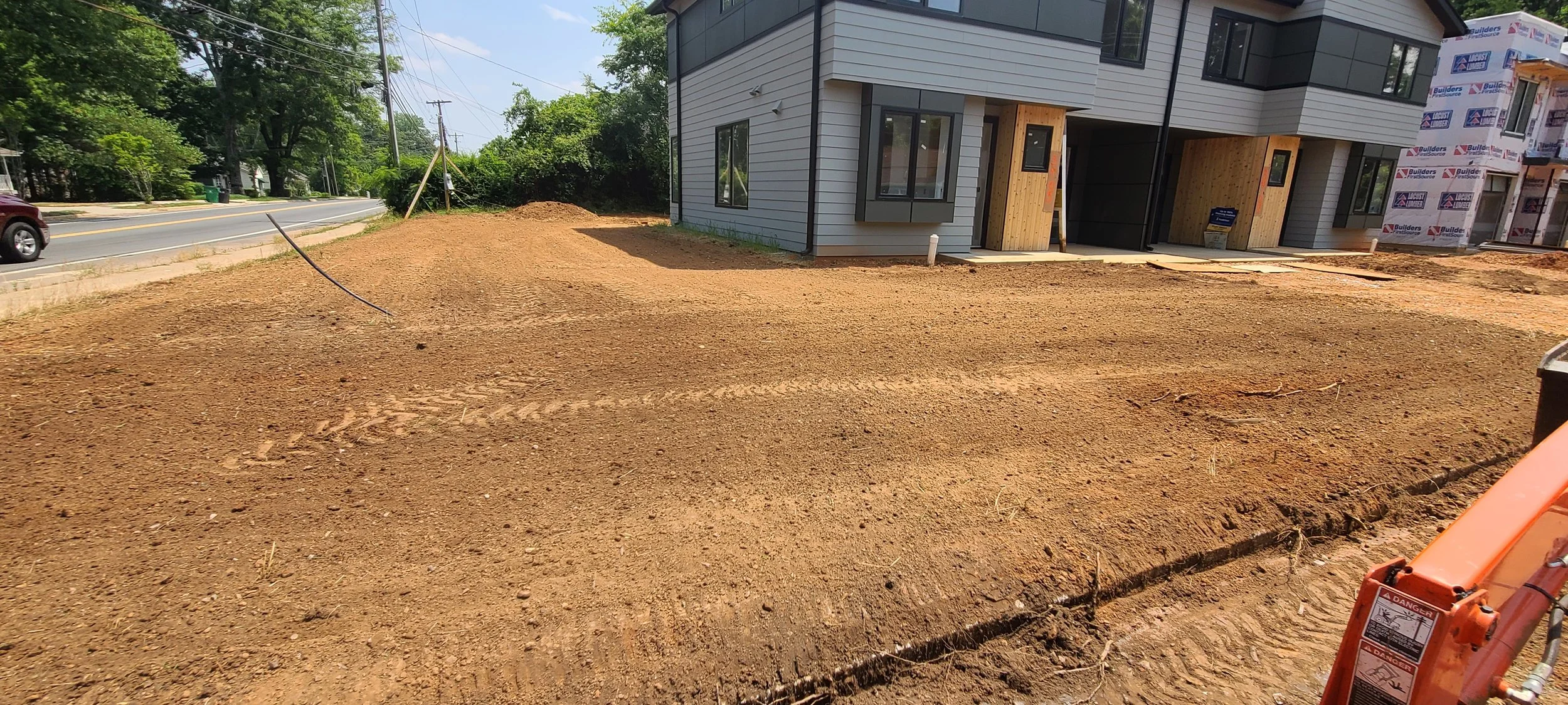 Construction site with disturbed dirt in front of a new multi-story residential building, partially built with siding and paper wrap, with windows and porch areas, set beside a paved road with a car passing by, and surrounded by trees.