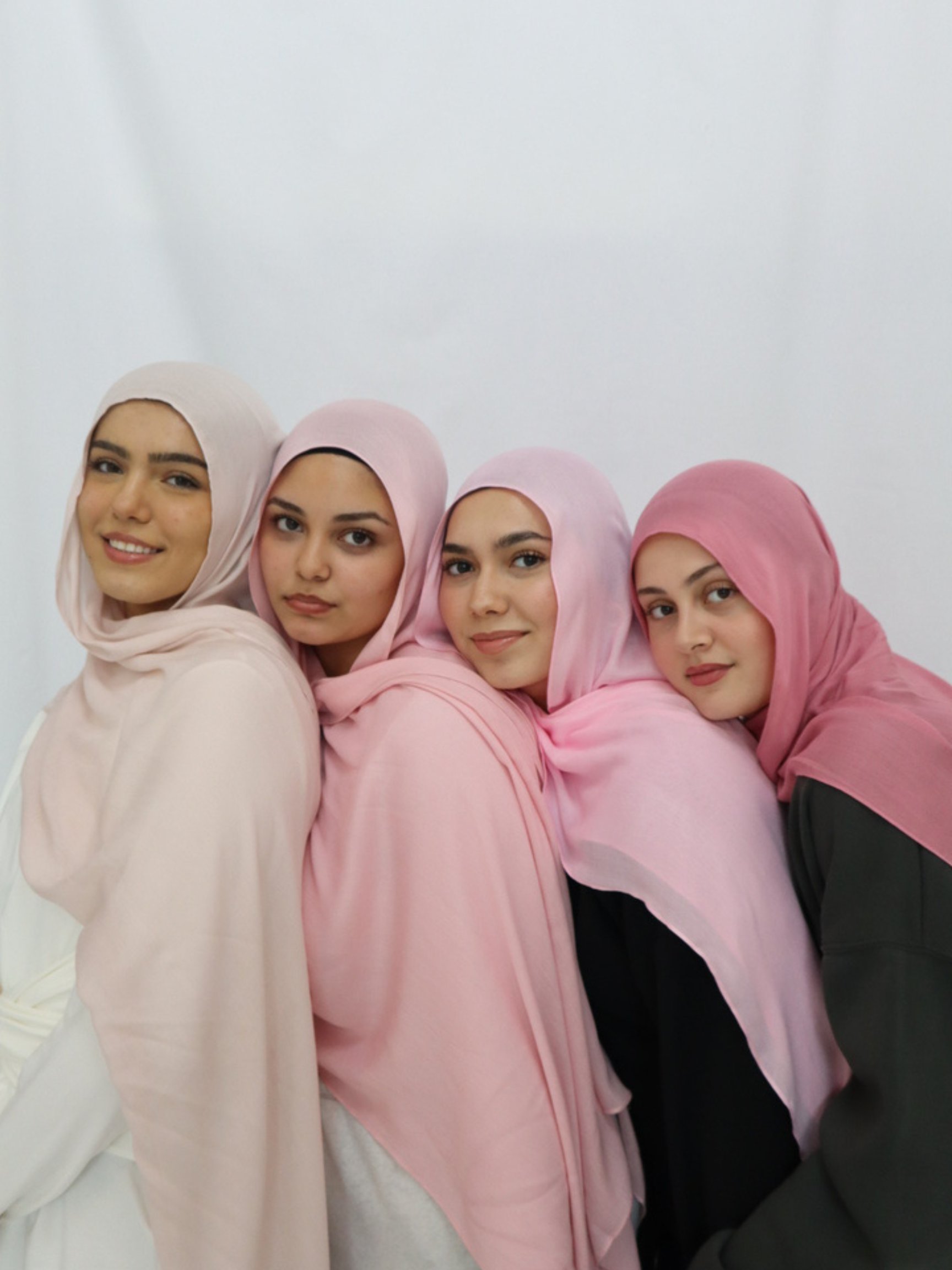 Four young women wearing hijabs in various shades of pink and beige, smiling and sitting close together against a white background.