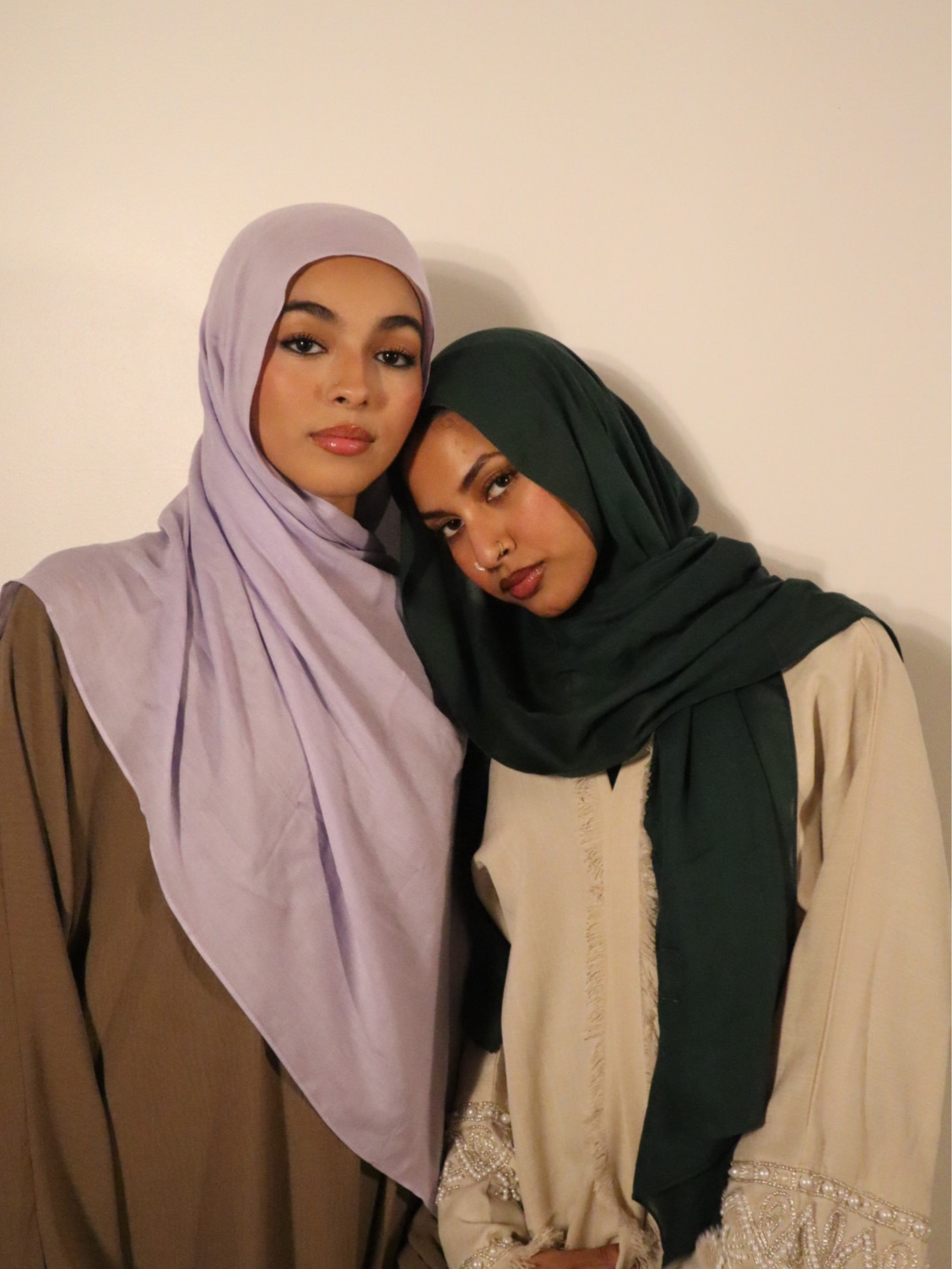 Two young women wearing hijabs, one in a lilac hijab and the other in a dark green hijab, standing close together against a plain wall.