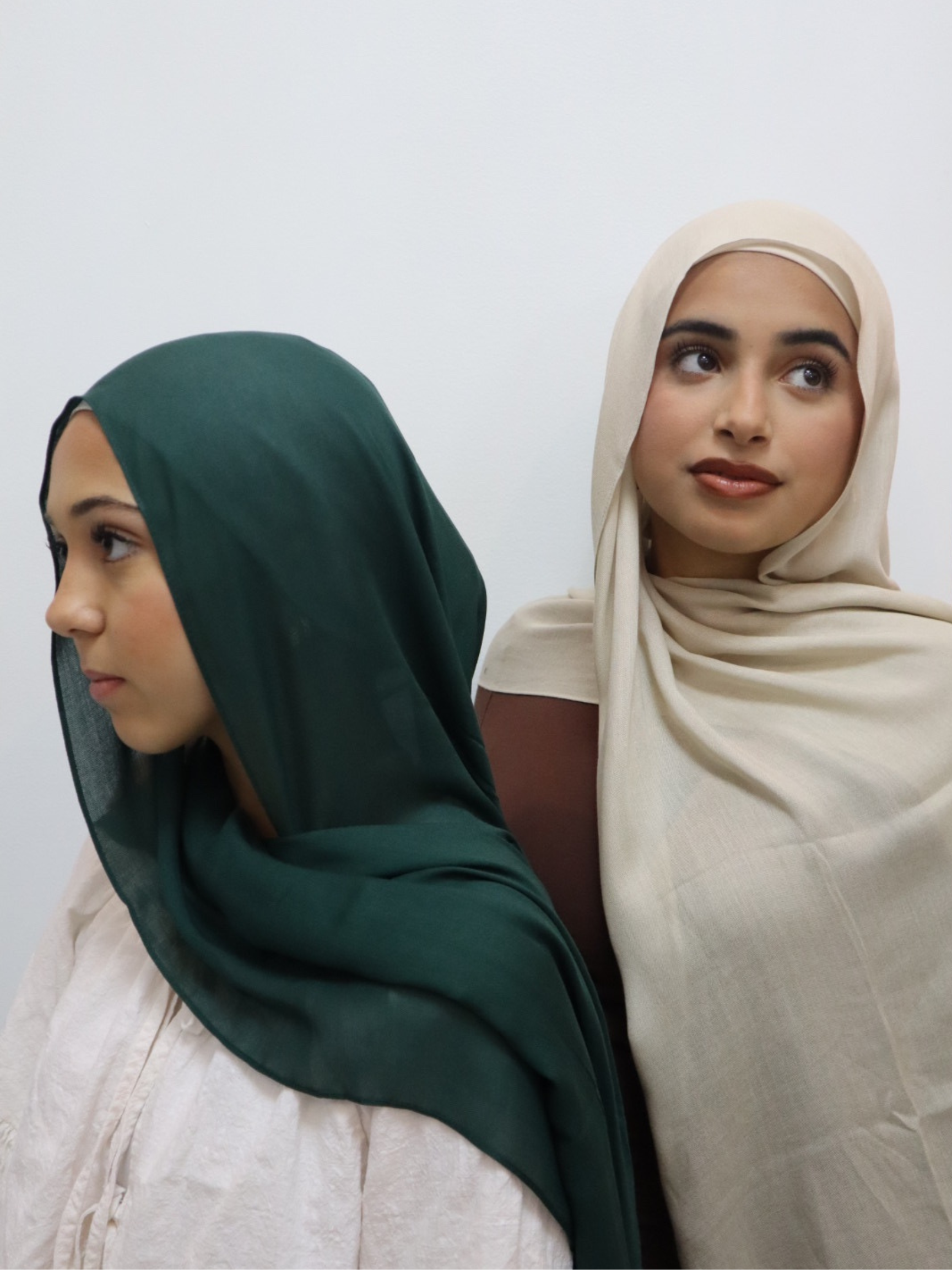 Two women standing side by side, wearing hijabs; one in dark green and the other in beige, against a plain white background.