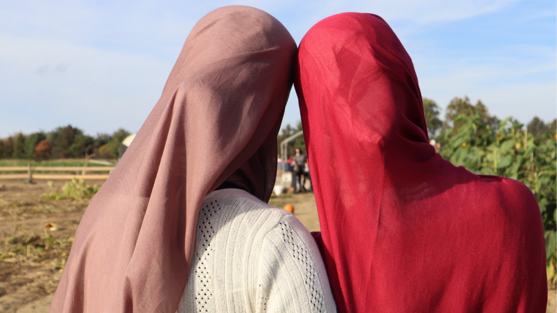 Two women with headscarves touching foreheads in a rural outdoor setting with farm fields and blue sky.